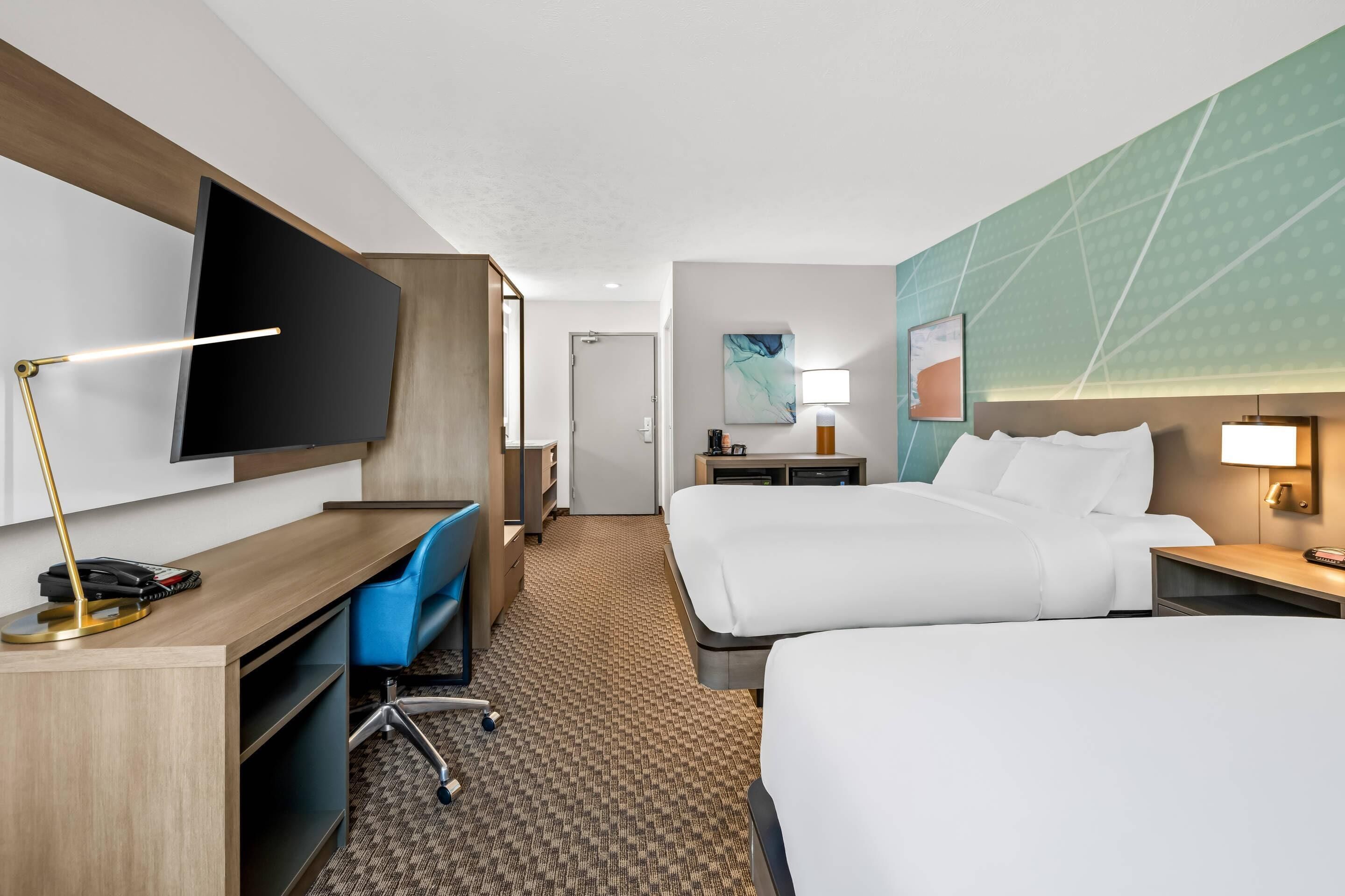 A hotel room with a desk, two beds, a TV, and a large window.