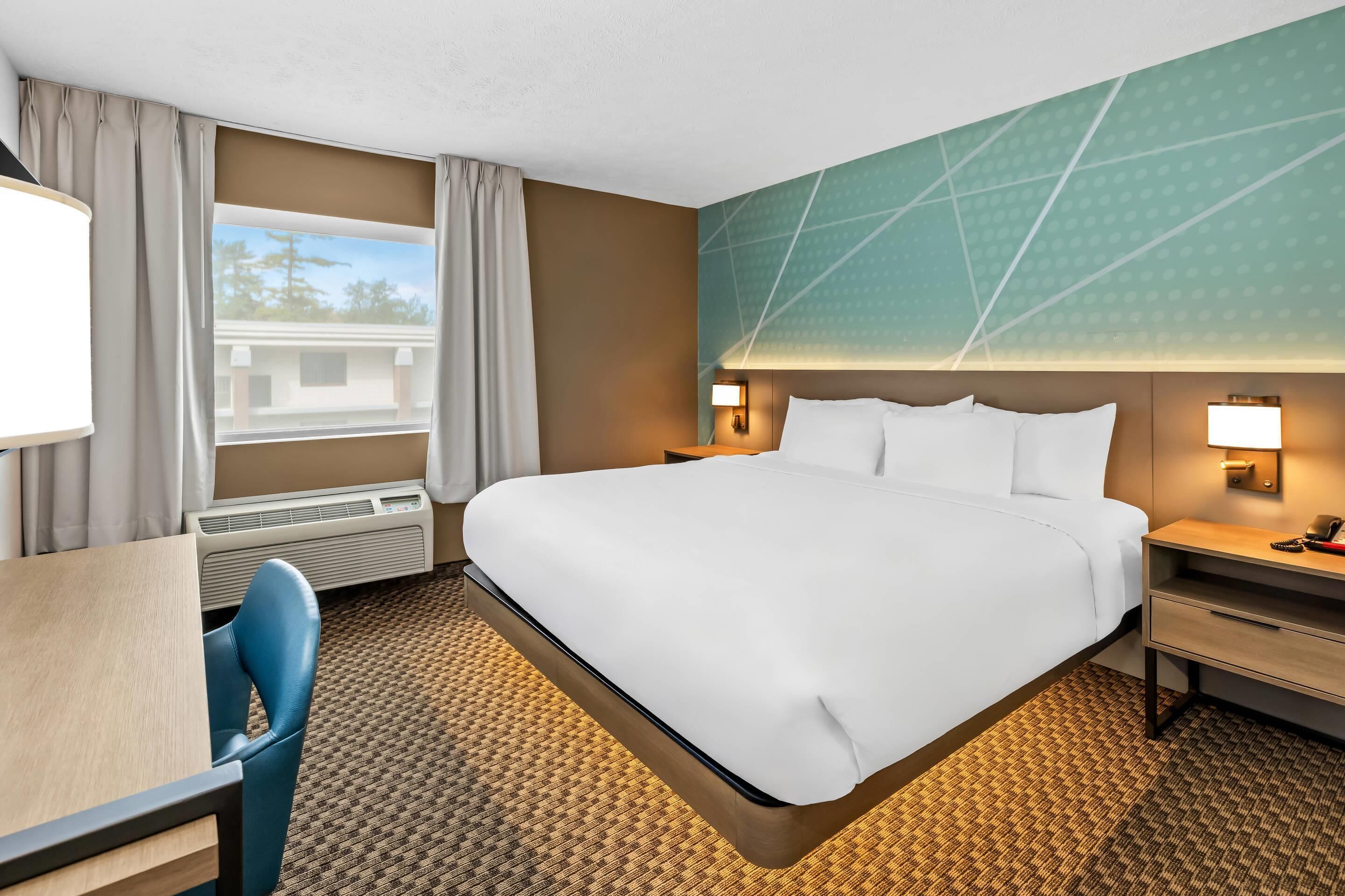 A hotel room with a large bed, a desk, a chair, a nightstand, and a wall-mounted lamp.