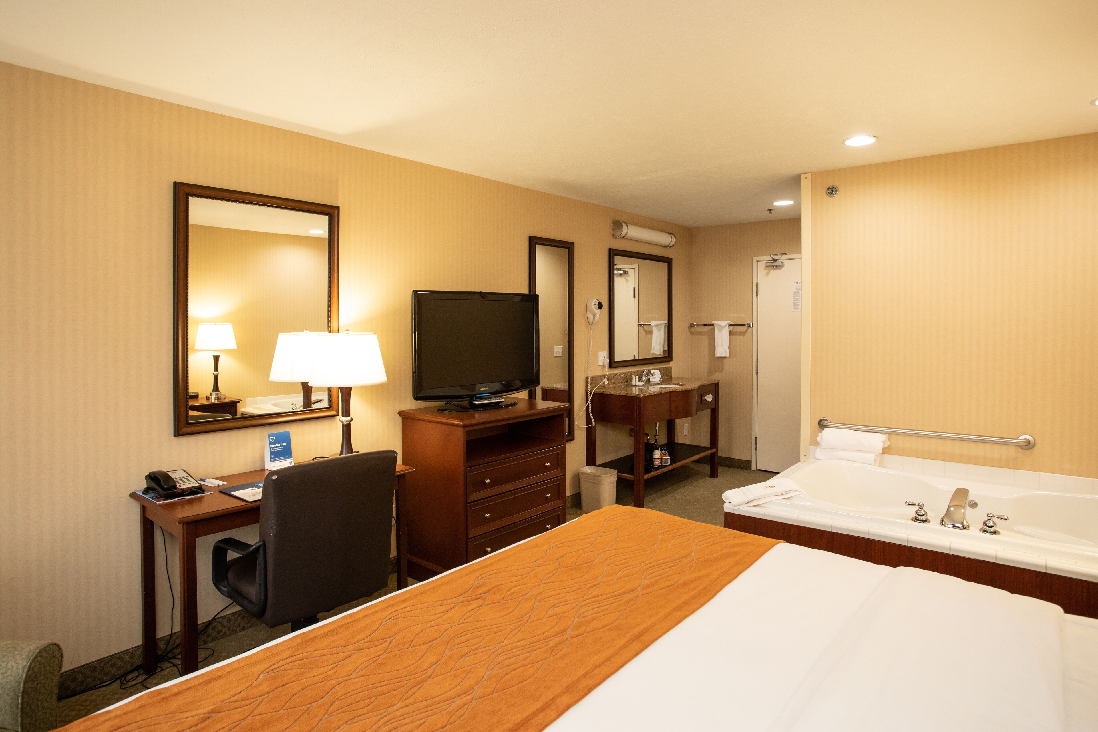 A hotel room with a bed, a desk with a computer, a television, a bathroom with a bathtub, and a mirror.