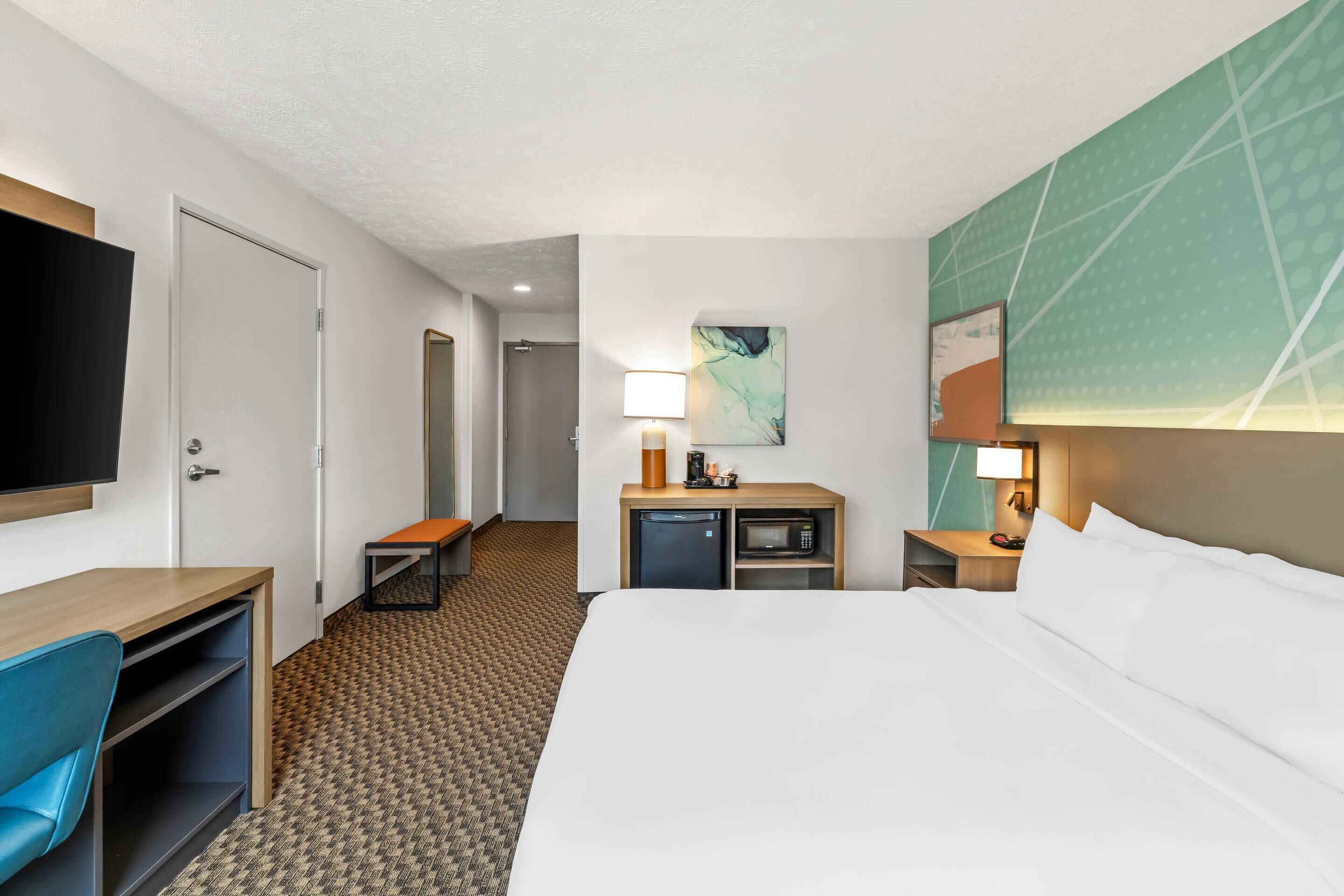 Habitación de hotel con cama, escritorio, silla, televisor, microondas y una mesita de noche con lámpara.