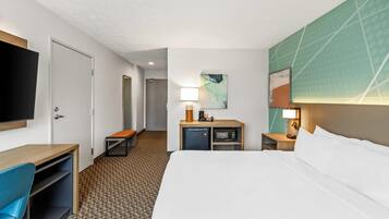 Habitación de hotel con cama, escritorio, silla, televisor, microondas y una mesita de noche con lámpara.