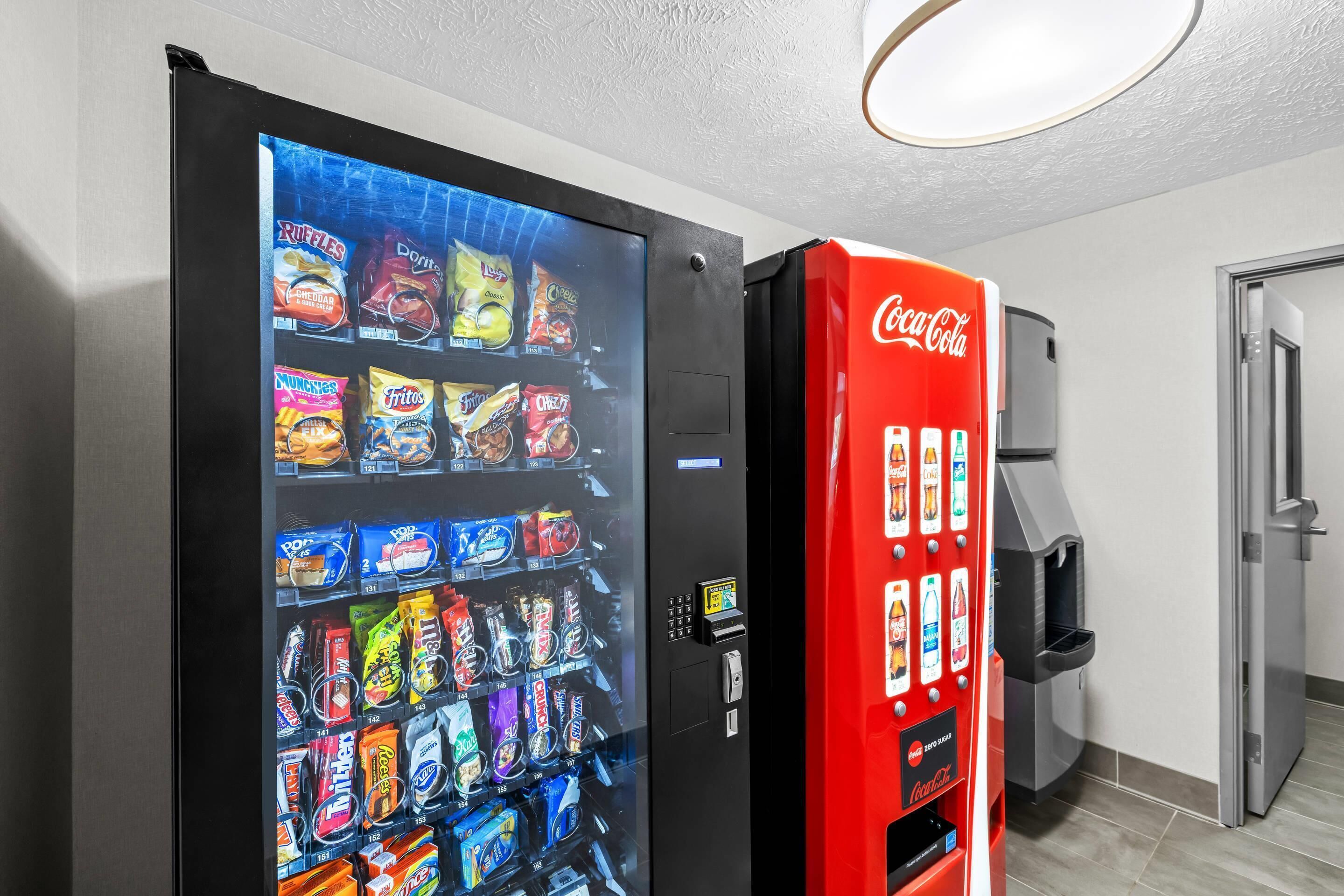 A vending machine with snacks and a Coca-Cola machine in a room with a door and a light fixture.