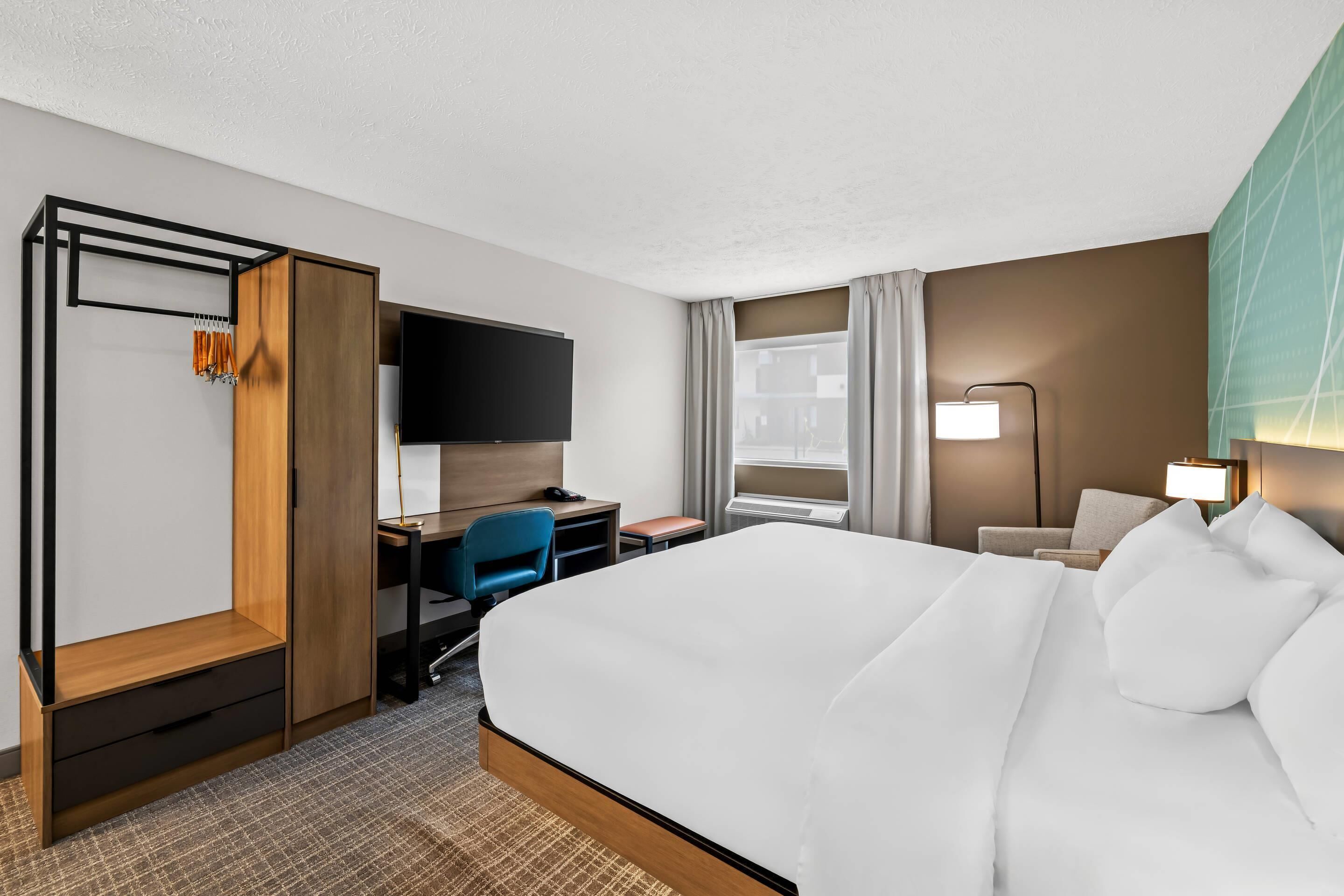 A hotel room with a large bed, a desk with a chair, a TV, a desk lamp, and a window with curtains.