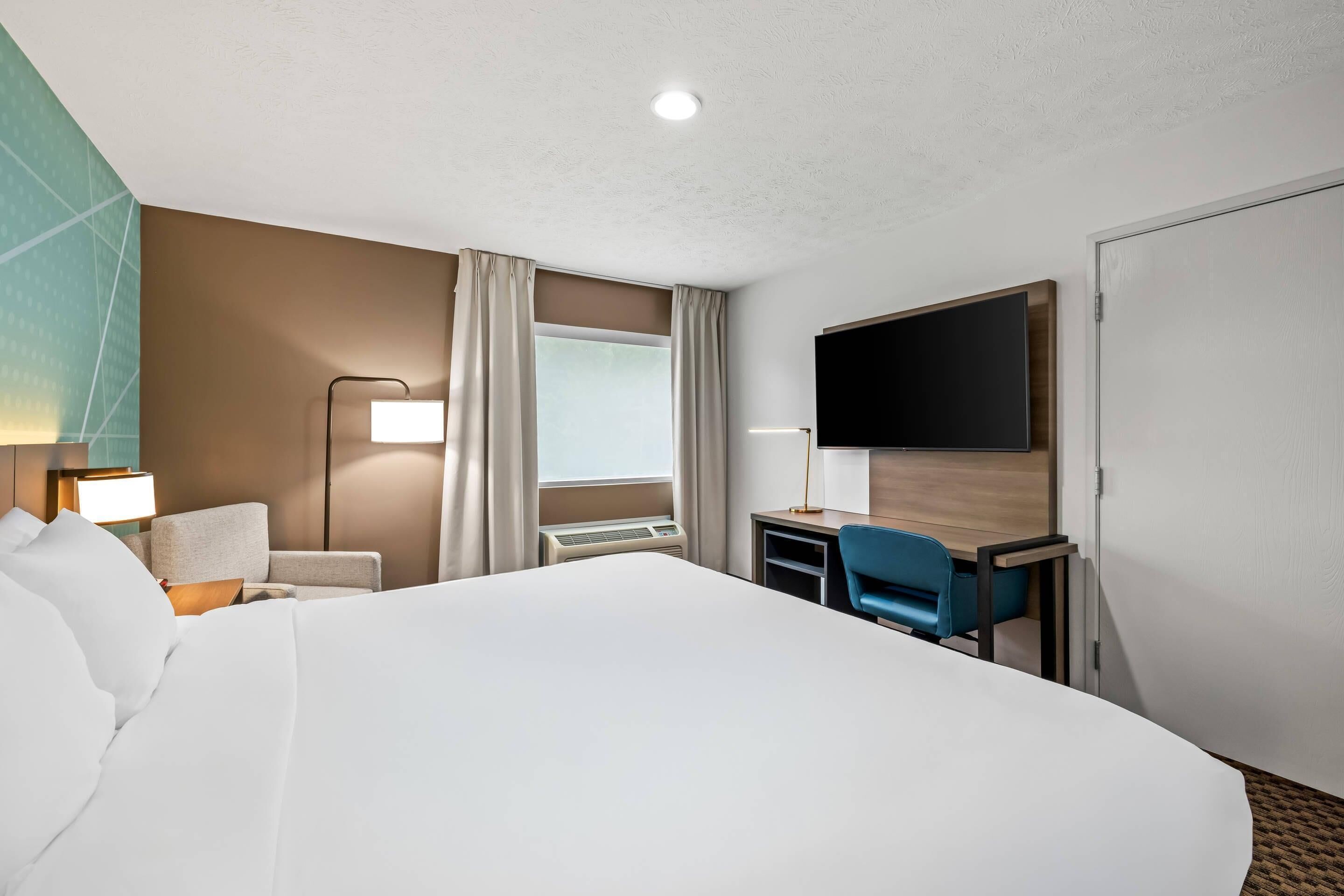 A hotel room with a large bed, a desk with a chair, a flat-screen TV, a lamp, and a window with curtains.