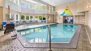 En inomhuspool med stora fönster som ger fri sikt mot himlen, omgiven av blå stolar och en möblerad sittgrupp under tak.