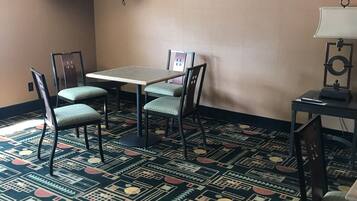A hotel room with a dining table, four chairs, a TV mounted on the wall, a framed picture, and a patterned carpet.