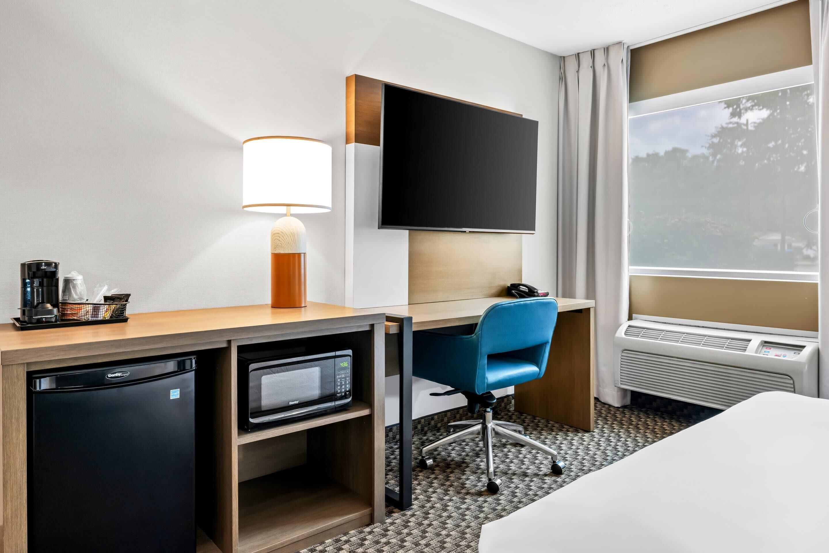 Une chambre d’hôtel équipée d’un bureau, d’une télévision à écran plat, d’un four à micro-ondes, d’un mini-réfrigérateur, d’une chaise bleue, d’un lit et d’une grande fenêtre avec des rideaux.