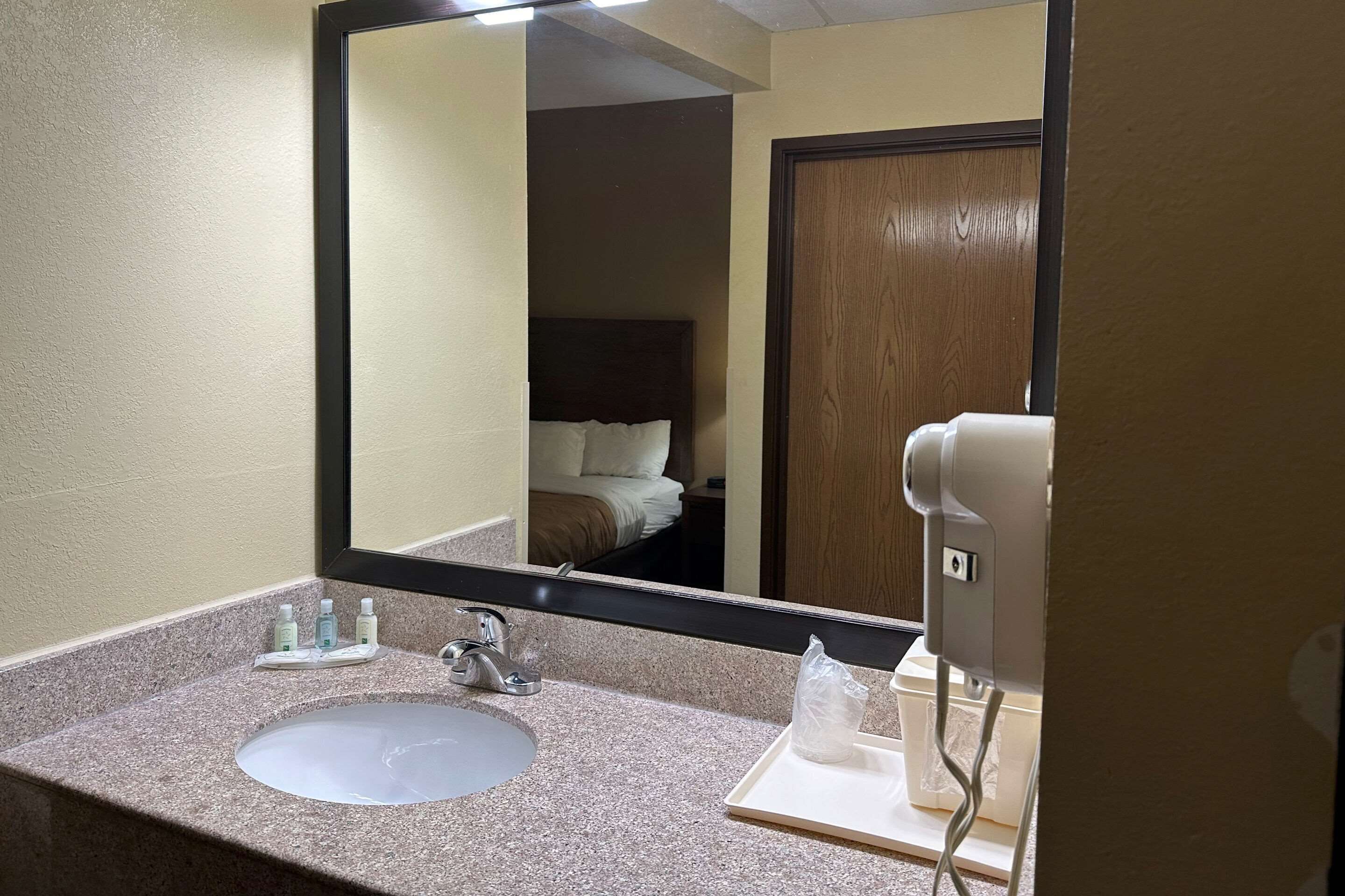 A hotel bathroom with a granite countertop, a sink, a mirror, and a hairdryer on a white stand.