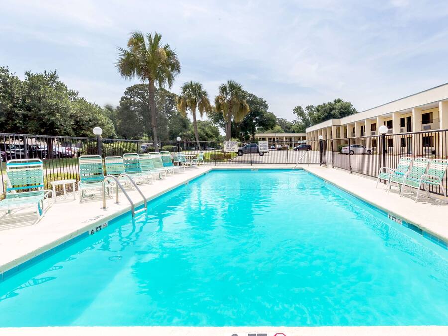 A swimming pool with lounge chairs and a fence, surrounded by palm trees and a building in the background.