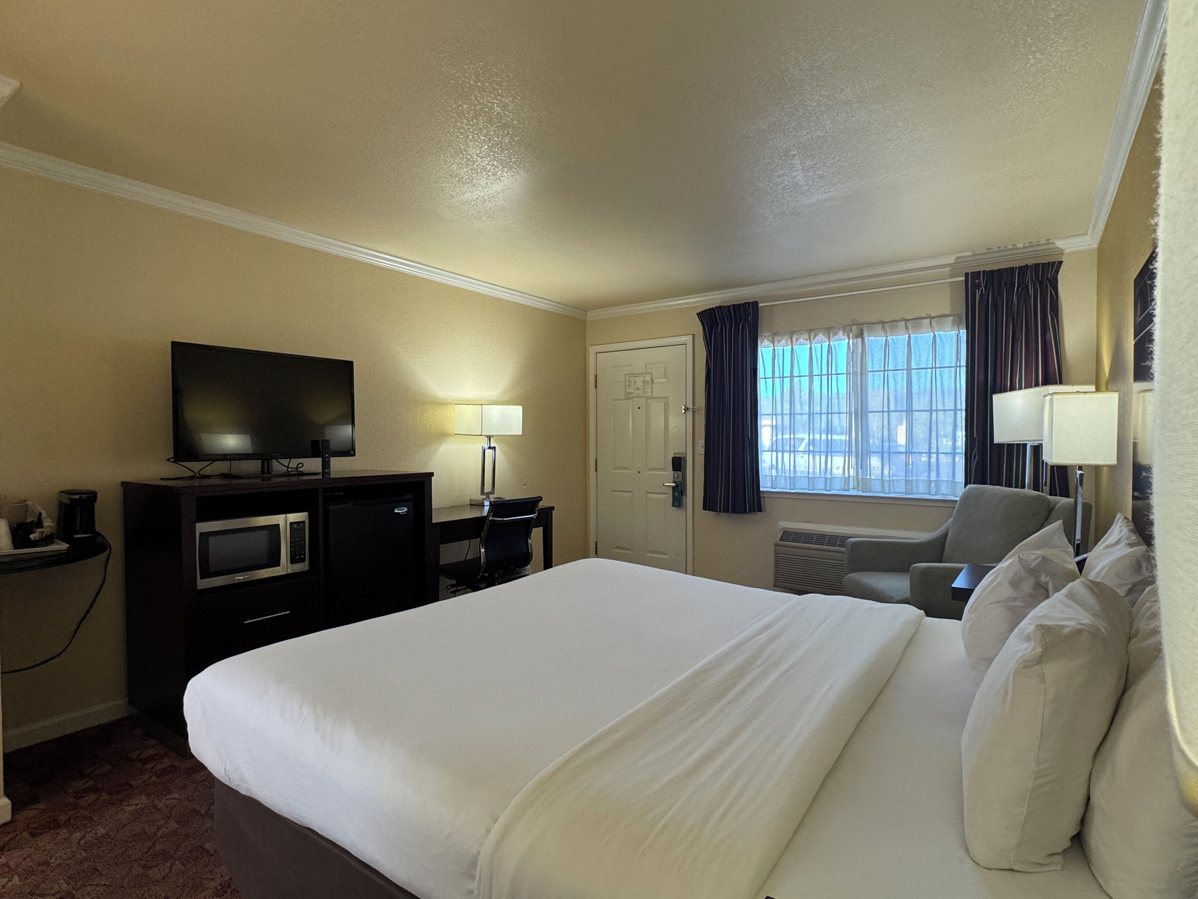 A hotel room with a large bed, a desk with a television, a microwave, and a coffee maker. There is a window with curtains and a chair.