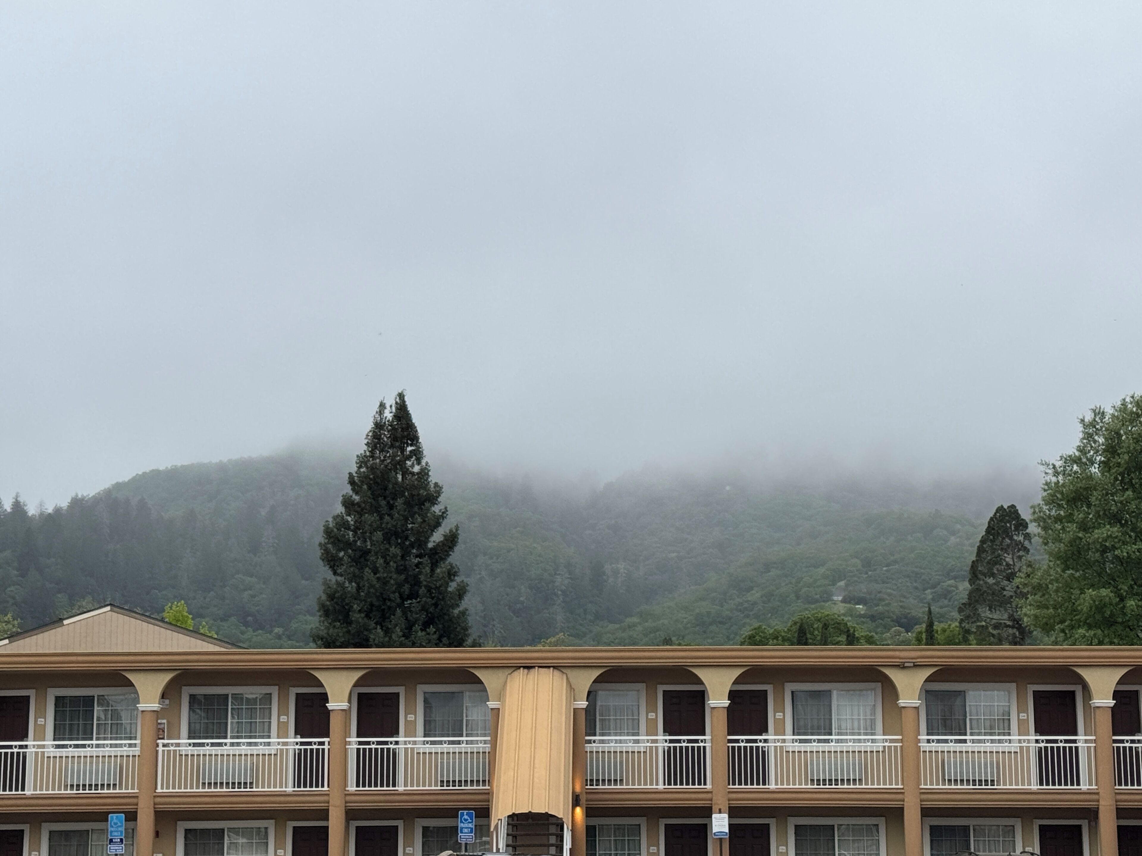 Een motel met meerdere verdiepingen, een balkon op de begane grond, een parkeersbord en een bosrijke heuvel op de achtergrond.