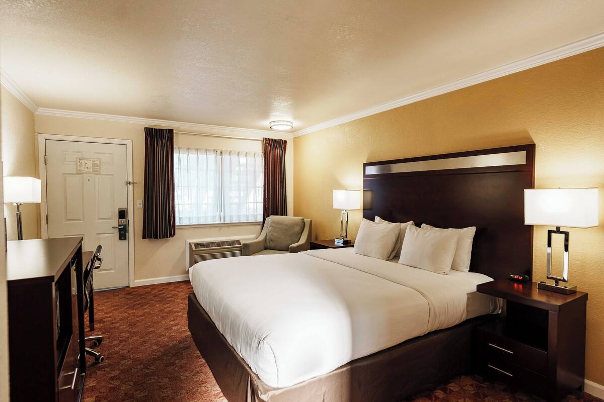 Ein Hotelzimmer mit einem großen Bett, Nachttischlampen, einem Schreibtisch mit Stuhl, einem Fernseher und einem Fenster mit Vorhängen.