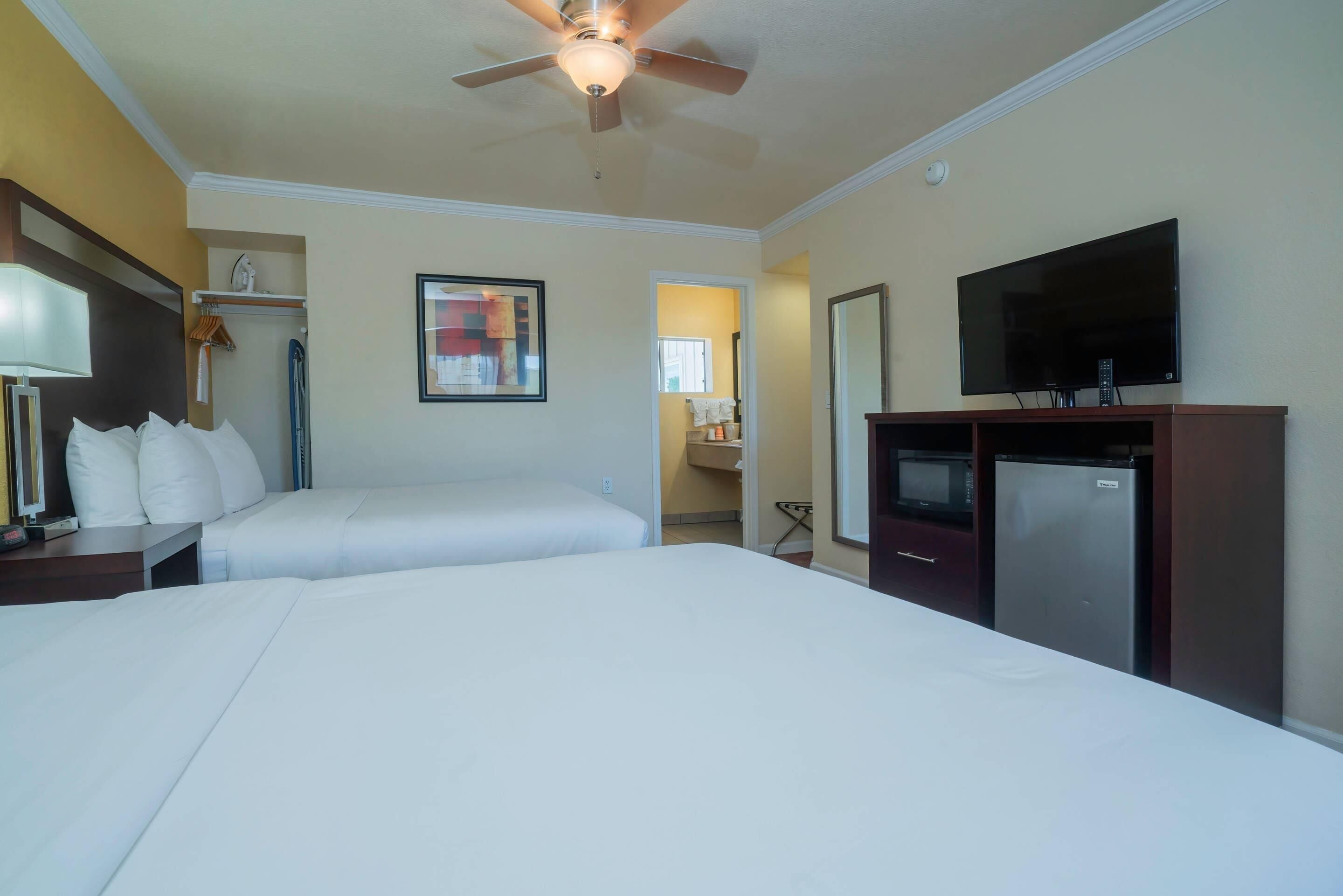 A hotel room with two beds, a ceiling fan, a TV, a refrigerator, and a bathroom visible through an open door.