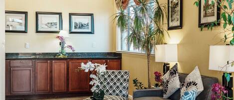 A spacious living area with a sofa, armchairs, a coffee table, and a dining table. There are framed pictures on the wall, a large window with curtains, and a potted plant.