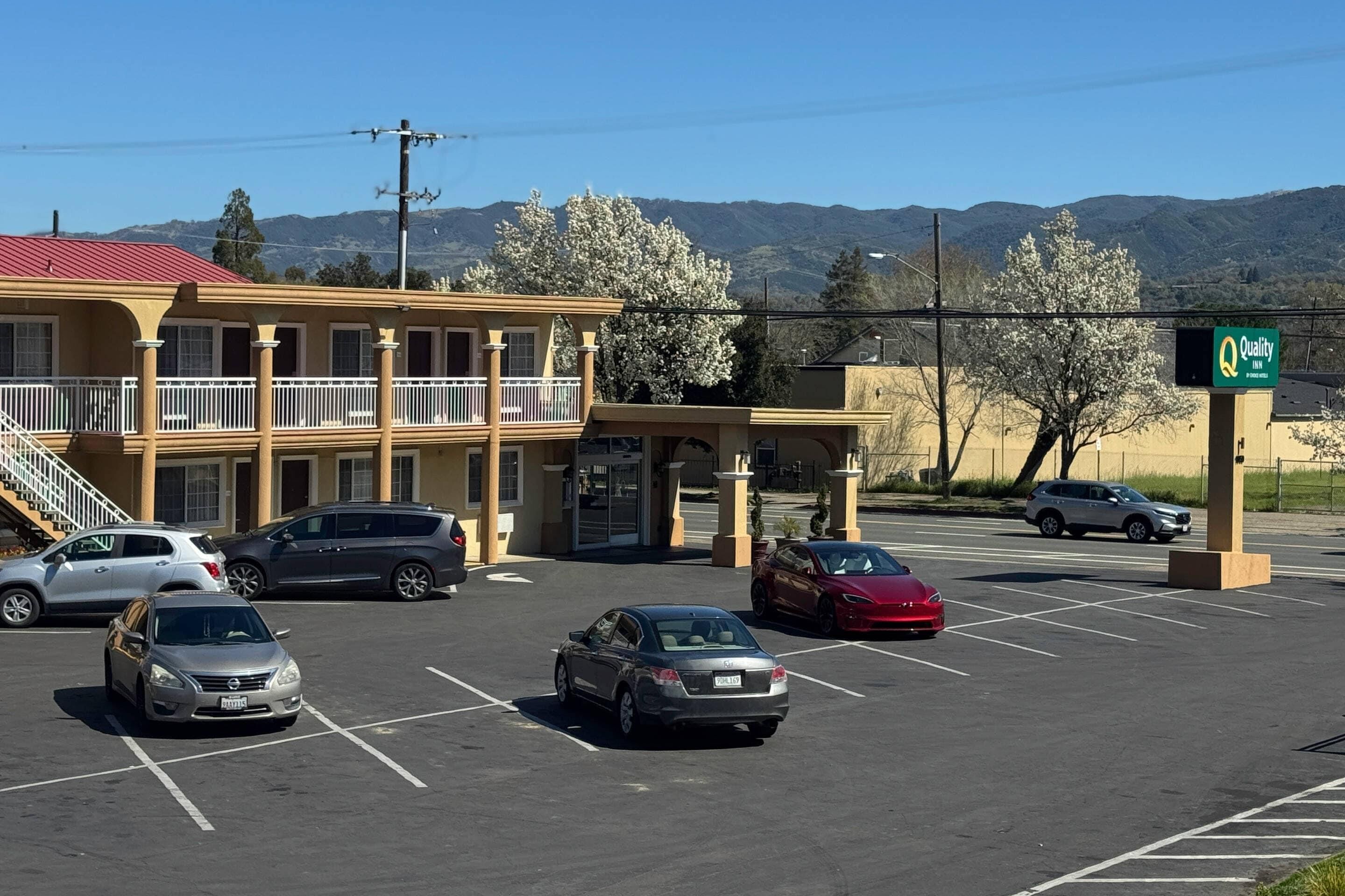 Un estacionamiento con varios autos, un edificio con balcón y montañas al fondo.