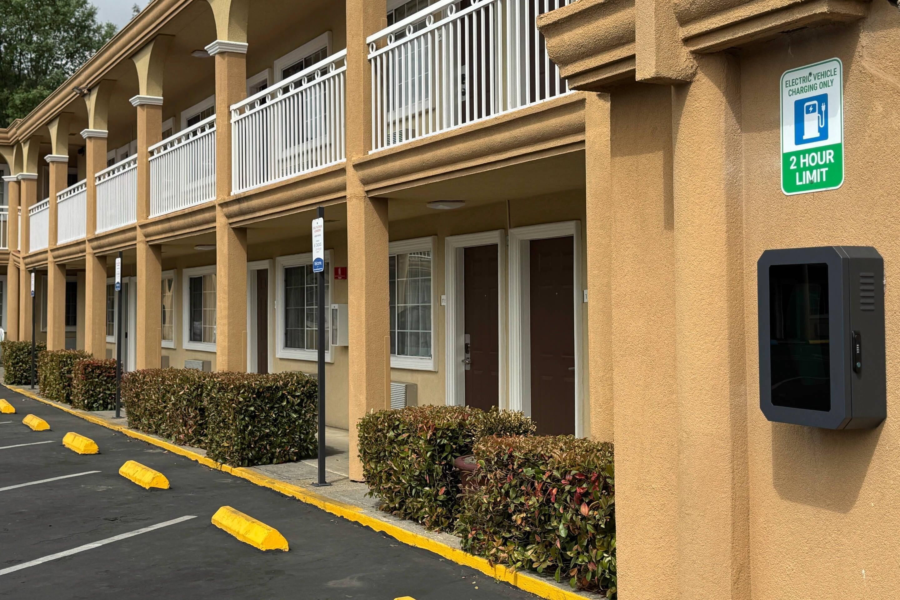 Un hotel con un cartel que indica un límite de 2 horas para la carga de vehículos eléctricos, estacionamiento exterior con barreras amarillas y una cámara de seguridad.