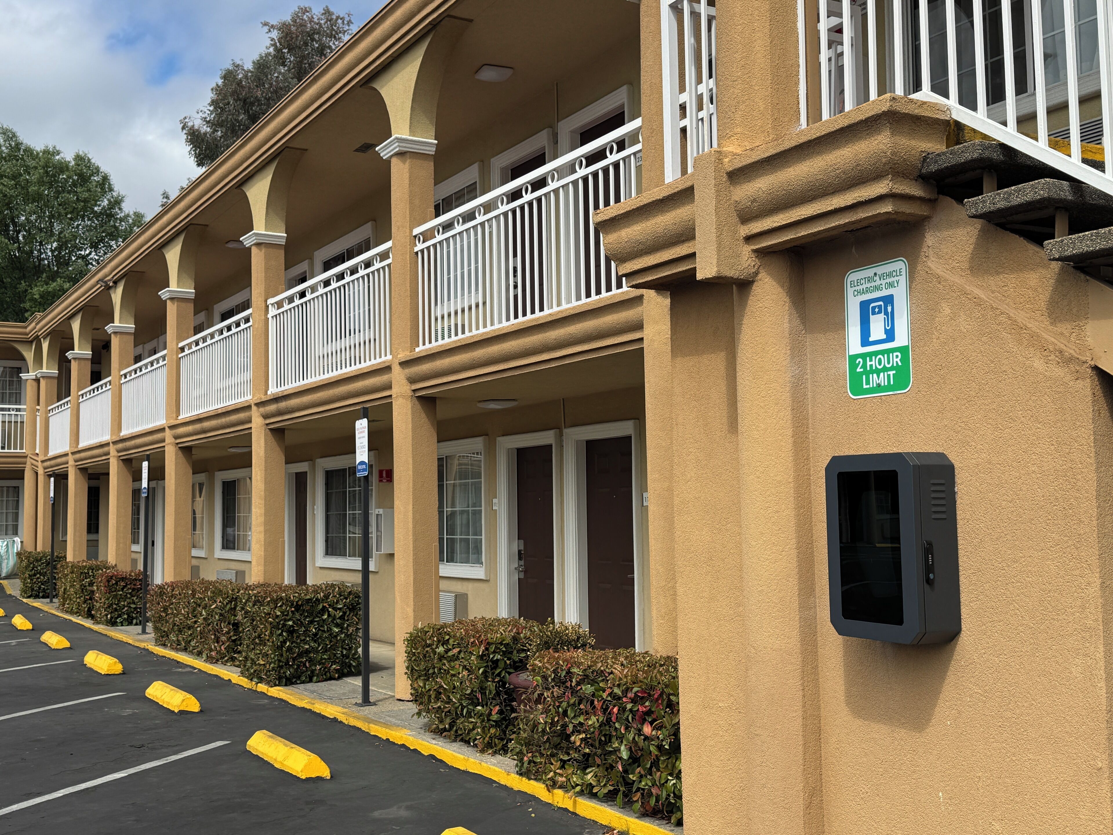 A hotel with a two-hour electric vehicle charging limit sign, a parking lot with yellow curbs, and a building facade with balconies.