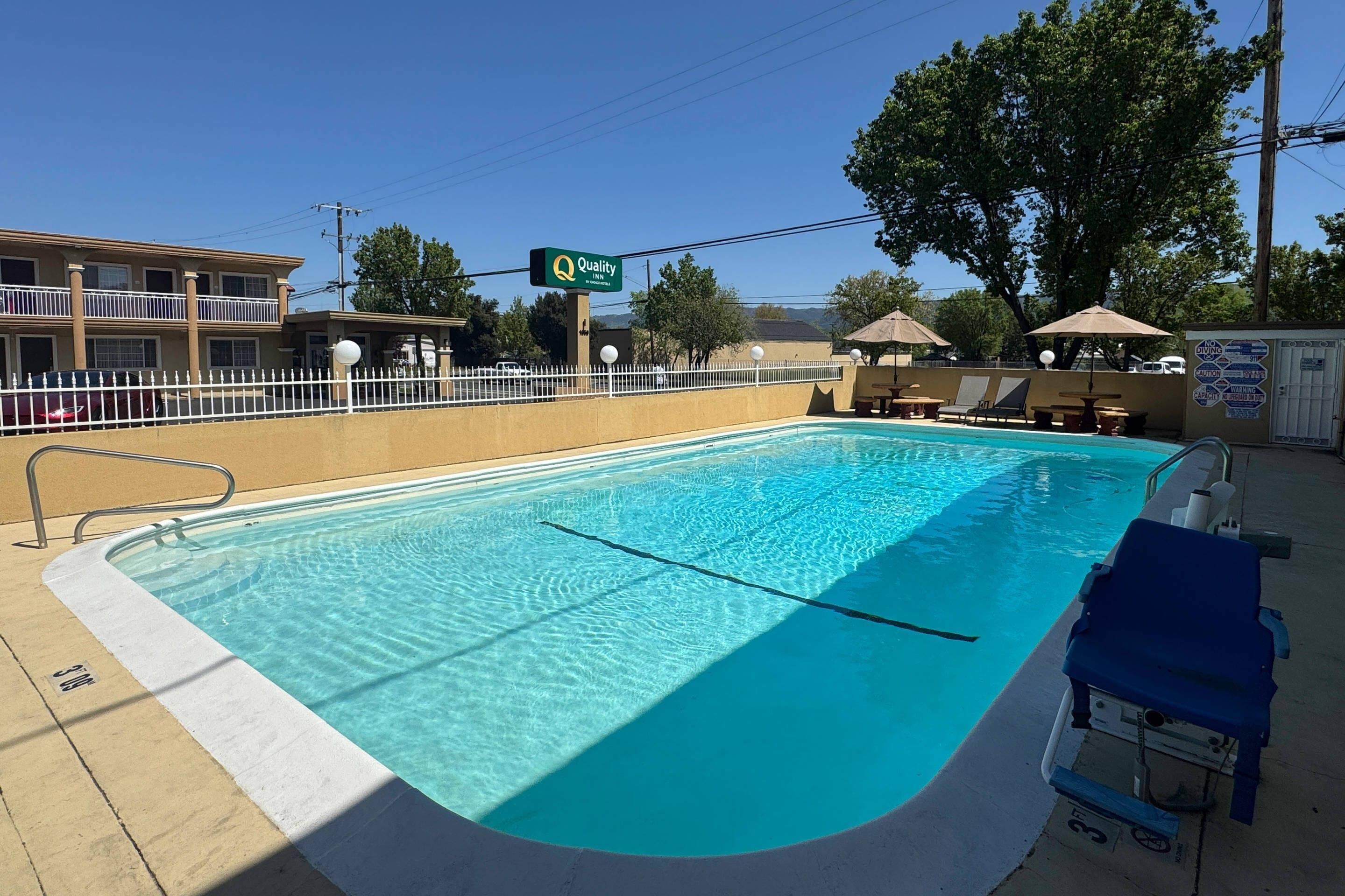 A swimming pool with a white deck, a sign reading "Quality", and a fenced area with lounge chairs.