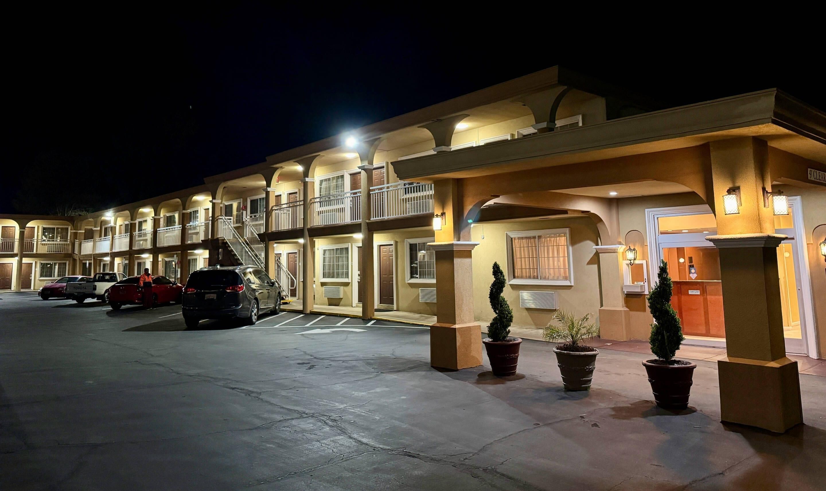 A multi-story hotel with a parking lot, featuring a central entrance with a staircase, potted plants, and a sign indicating a clear area.