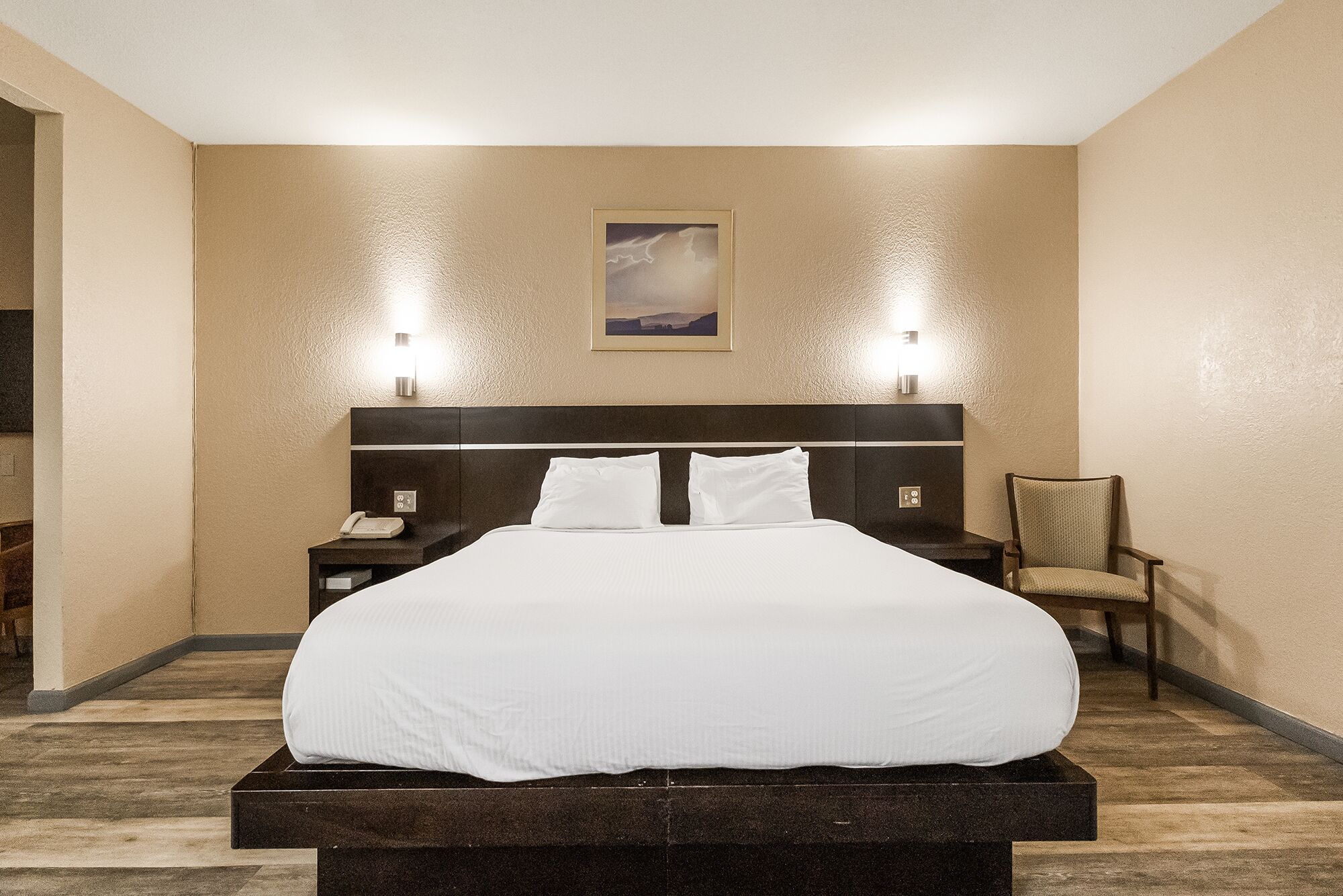 A hotel room with a large bed, two bedside lamps, a chair, and a painting on the wall.