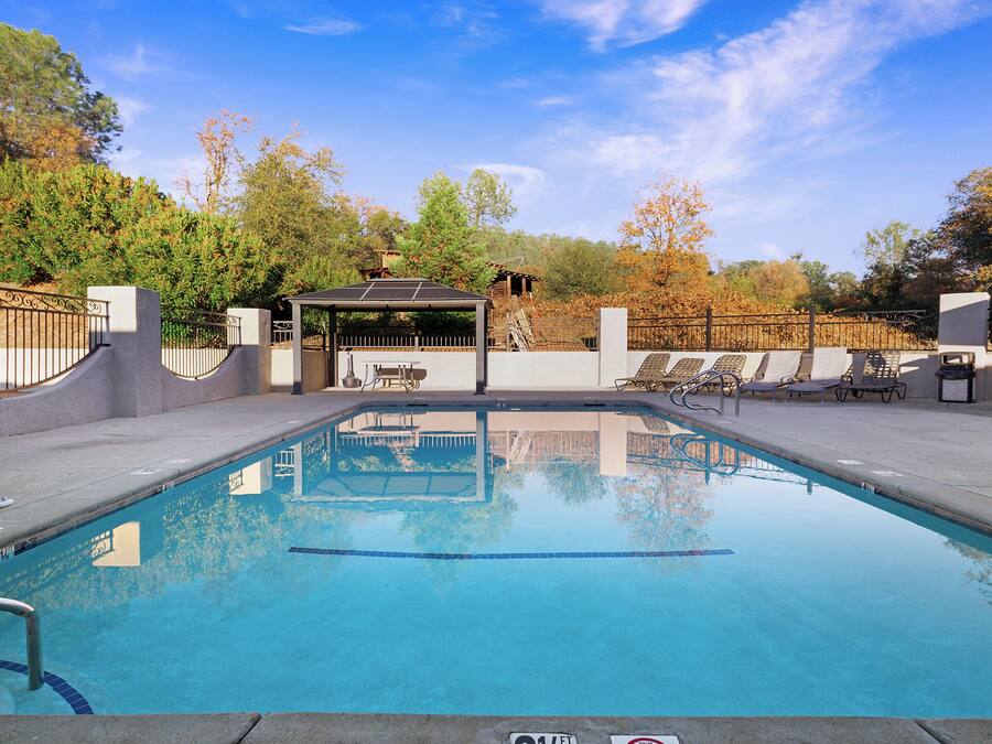 A clear swimming pool with lounge chairs and a covered seating area, surrounded by trees and a fence.