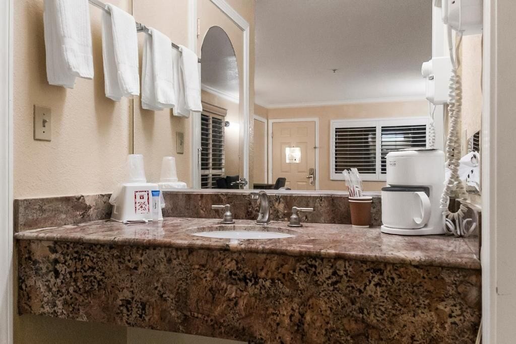 A bathroom with a granite countertop, a sink, a mirror, a towel rack, a coffee maker, and a telephone.