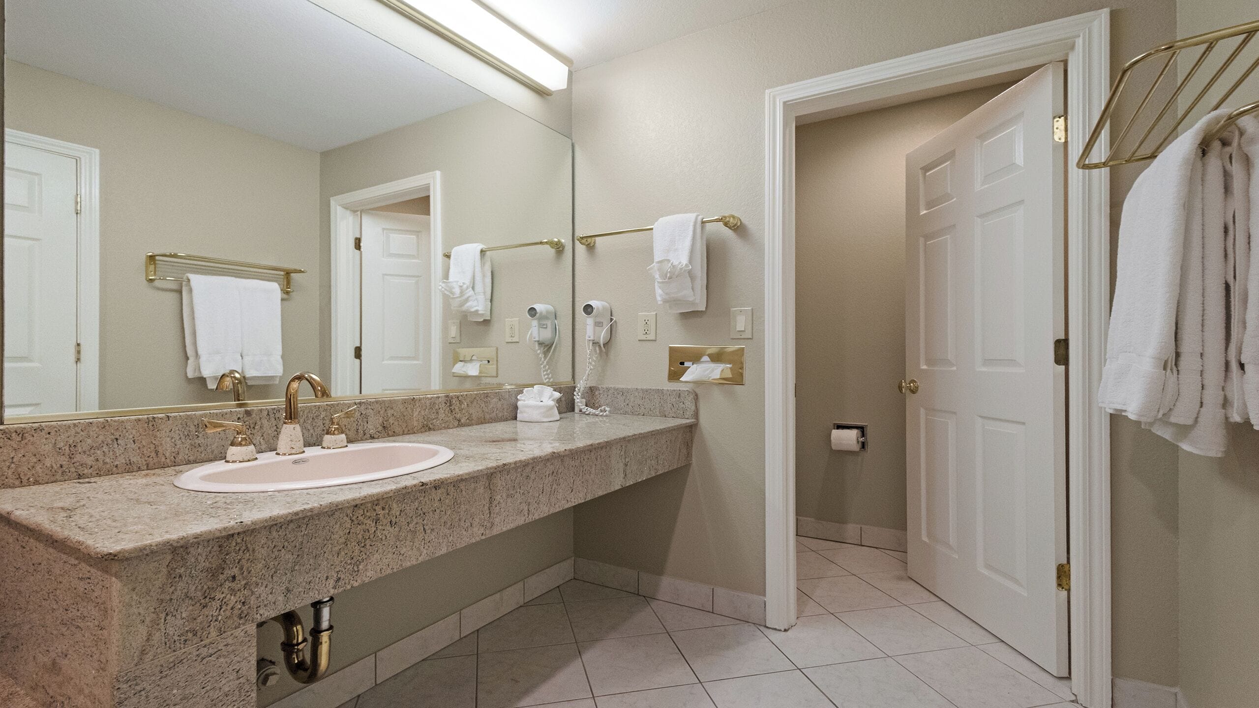 Un baño con encimera de granito, un lavabo, un espejo, un perchero para toallas y una puerta que da a otra habitación.