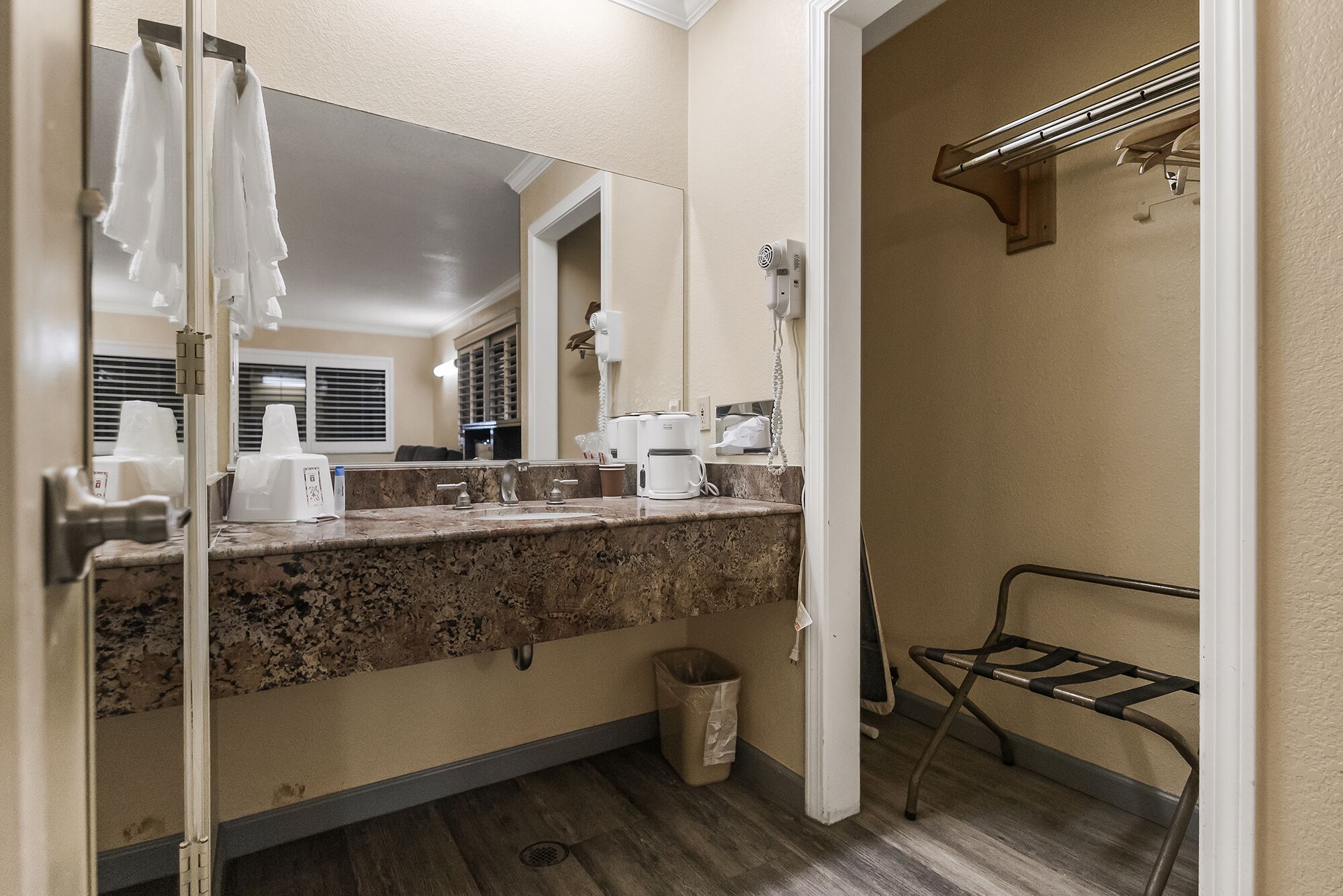 A bathroom with a granite countertop, a mirror, a towel rack, a coffee maker, and a trash can.
