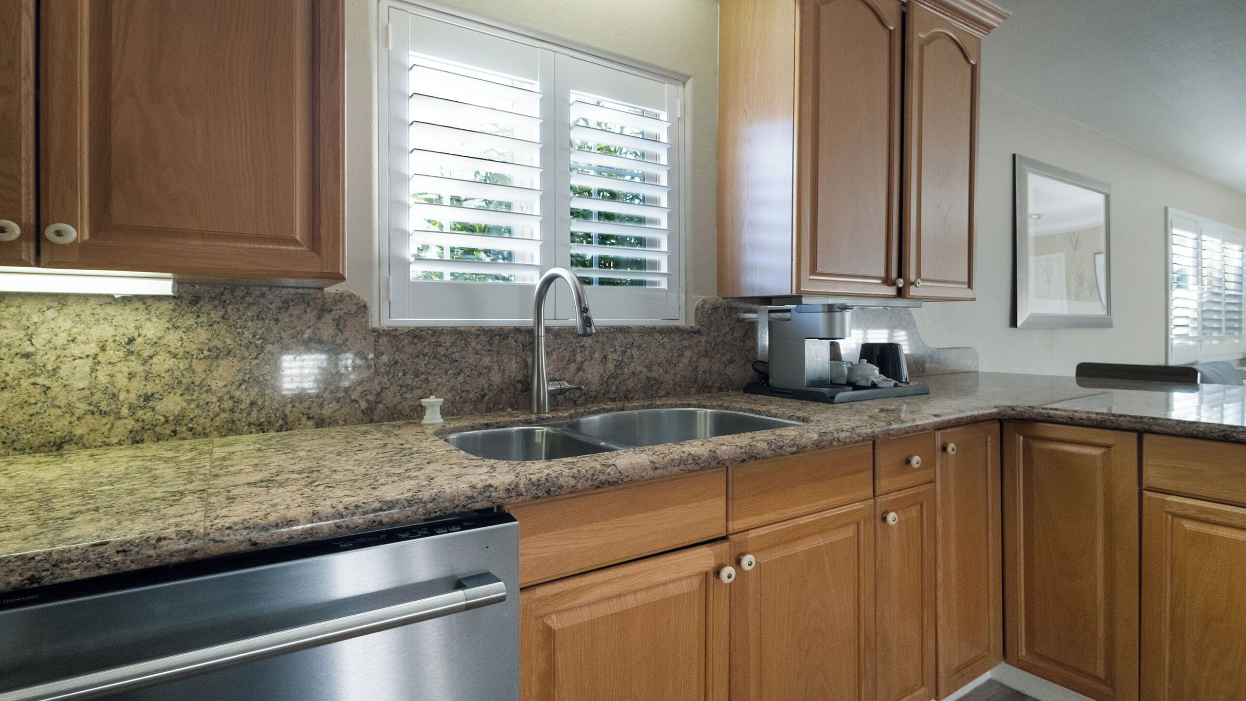 Una cocina con encimeras de granito, electrodomésticos de acero inoxidable, gabinetes de madera y un ventanal con persianas.