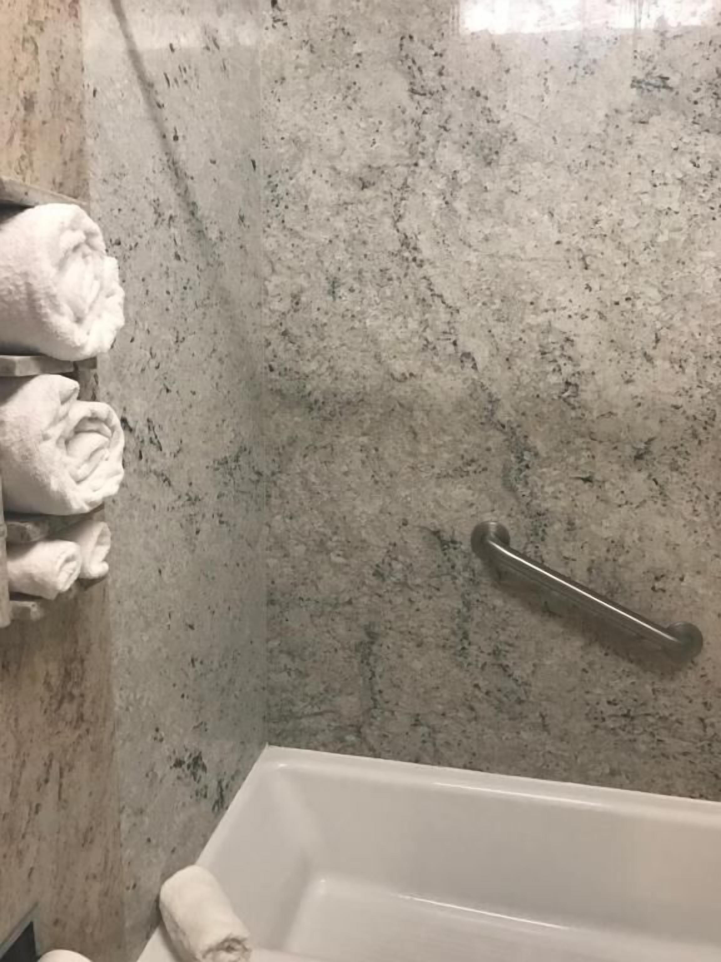 A bathroom with a marble wall, a towel rack with folded towels, and a grab bar near the bathtub.
