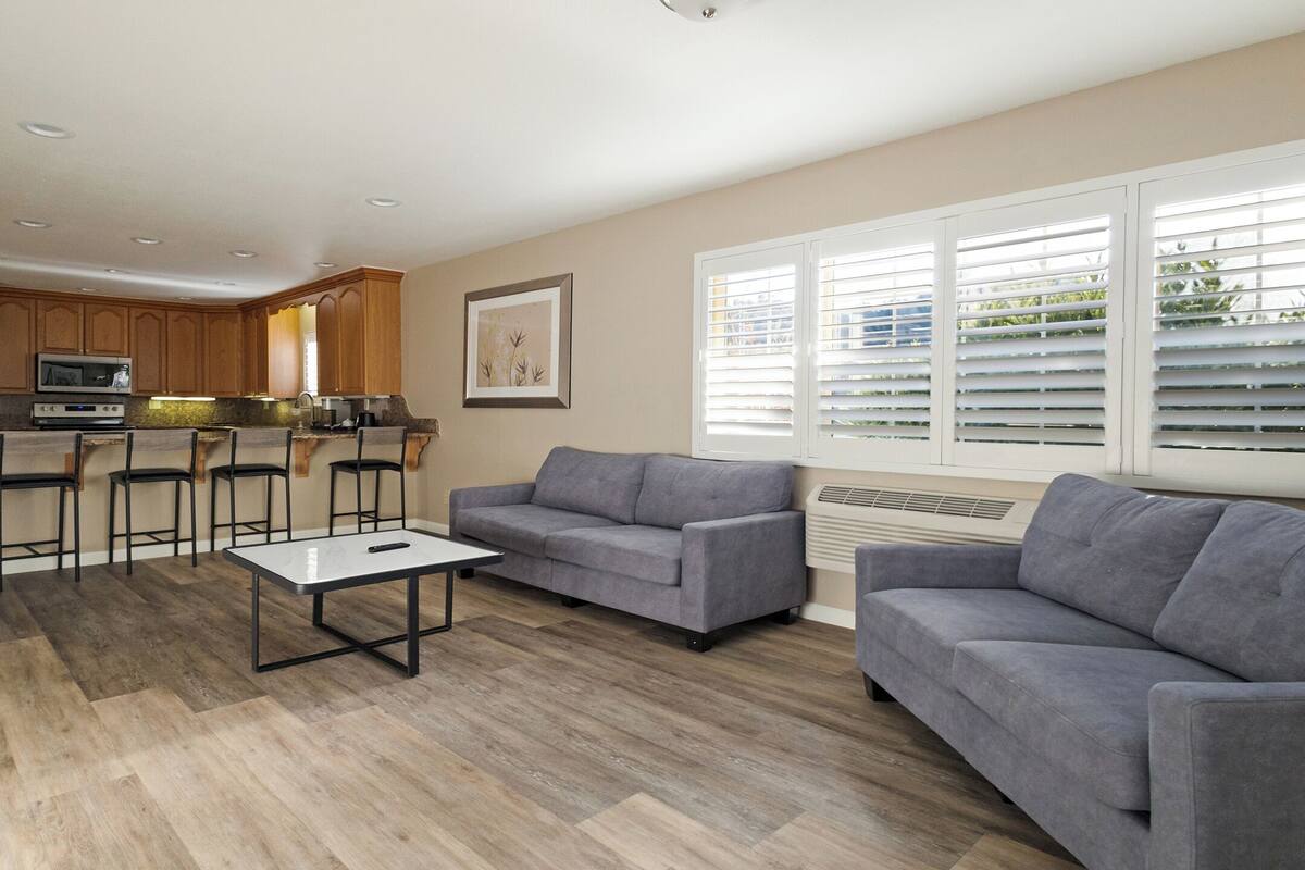 A modern living room with a grey sofa, a coffee table, and a kitchen area with wooden cabinets and a built-in oven.