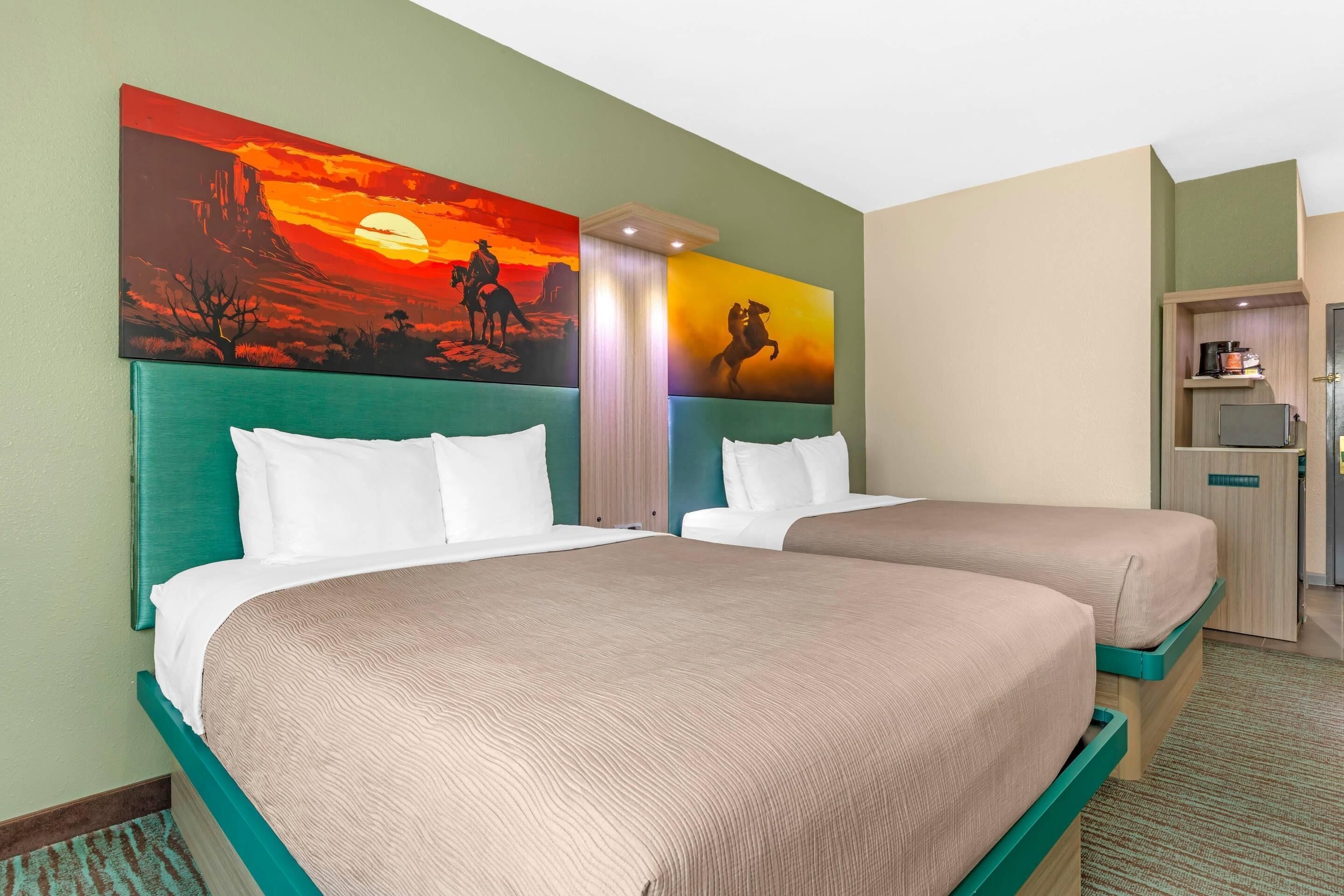 A hotel room with two beds, a large painting on the wall, a small kitchenette, and a carpeted floor.