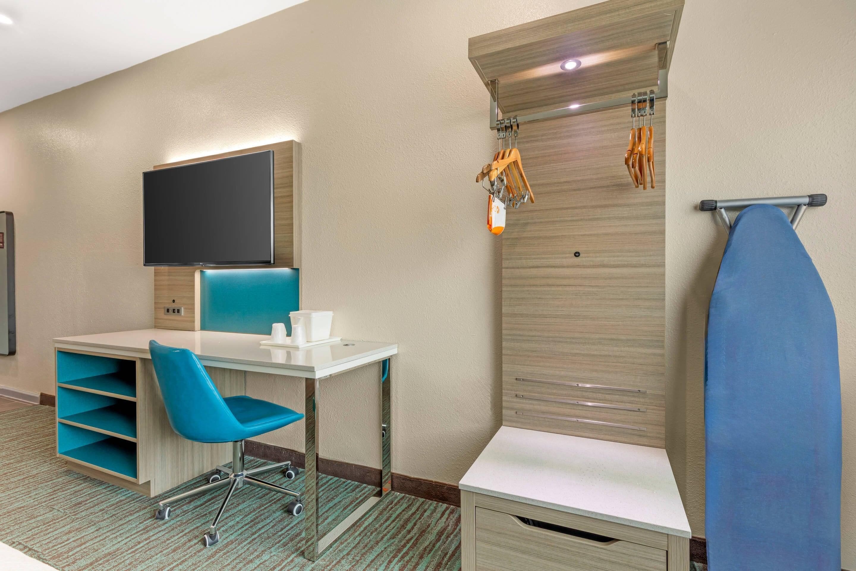 A hotel room with a desk, chair, television, ironing board, and storage shelves.