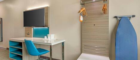 A hotel room with a desk, chair, television, ironing board, and storage shelves.