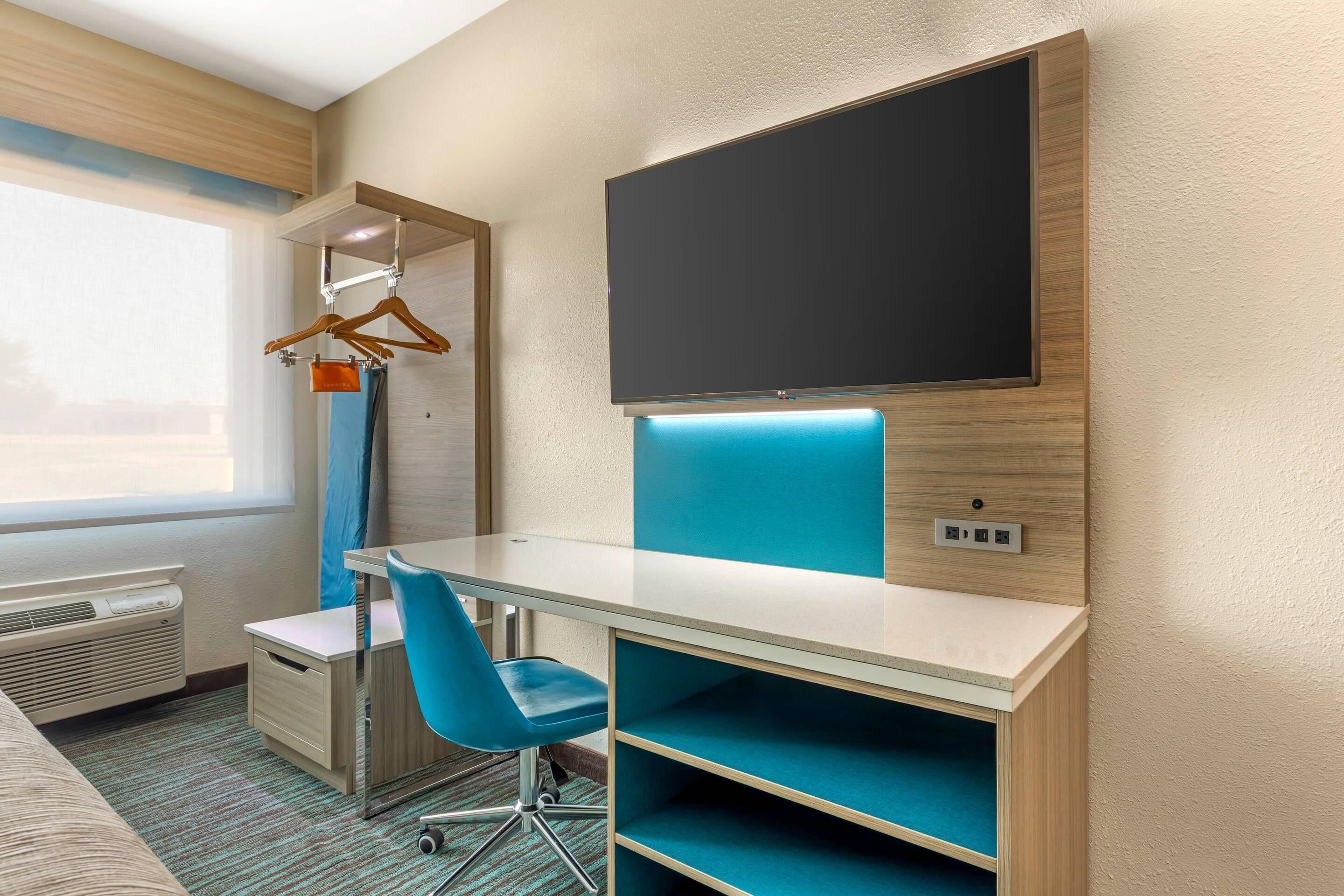 A hotel room with a desk, chair, TV, and a coat rack.