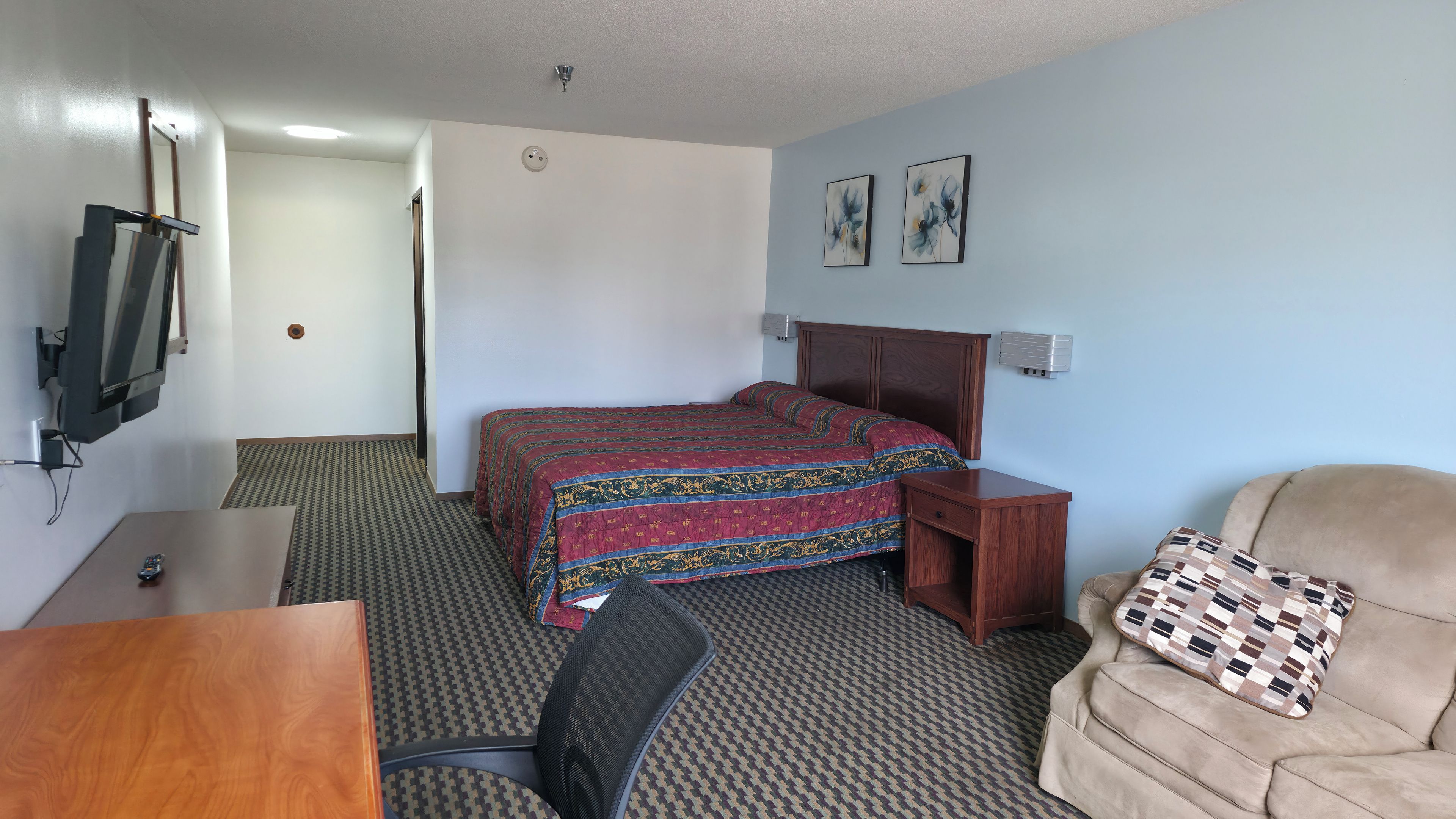 A hotel room with a bed, a desk, a chair, a television, and a wall-mounted air conditioning unit.