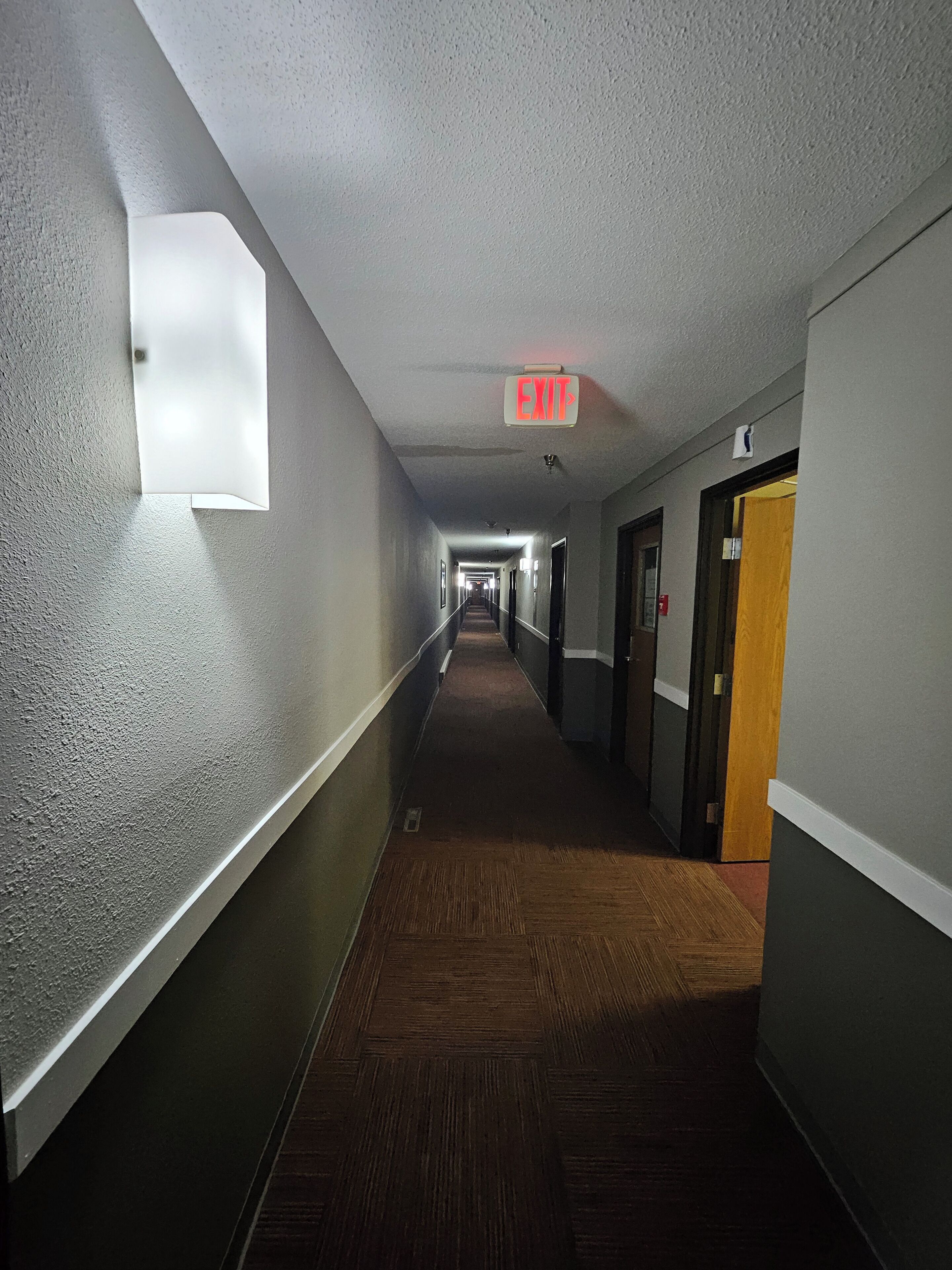 A long, narrow hotel corridor with a carpeted floor, a light fixture on the left wall, and an exit sign on the ceiling.