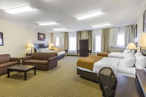 A hotel room with a large bed, two sofas, a small table, and a desk. There are curtains on the windows, a door, and a flat-screen TV on the wall.