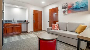 A modern living room with a grey sofa, a red ottoman, and a kitchen area with wooden cabinets and a black countertop. - Comfort Inn Livingston (Livingston)