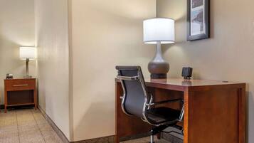 Habitación de hotel con un escritorio de madera, una silla de oficina negra, una lámpara, una mesita con un teléfono y un cuadro en la pared.