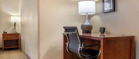 Habitación de hotel con un escritorio de madera, una silla de oficina negra, una lámpara, una mesita con un teléfono y un cuadro en la pared.