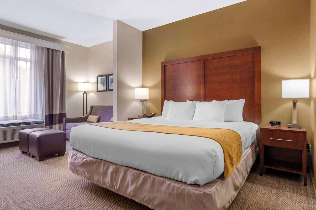 Ein Hotelzimmer mit einem großen Bett, einem Nachttisch mit Lampe, einem Sessel und einem Fenster mit Vorhängen.