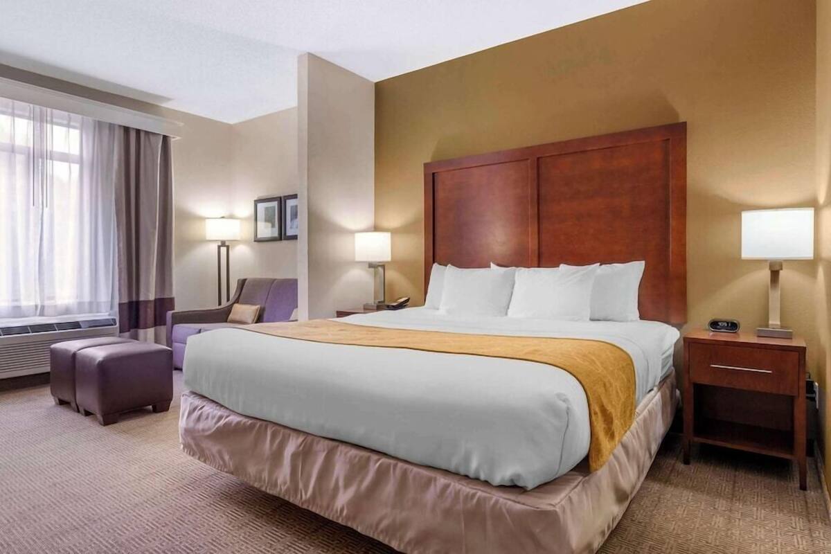 Ein Hotelzimmer mit einem großen Bett, einem Nachttisch, einer Lampe, einem Sessel und einem Fenster mit Vorhängen.