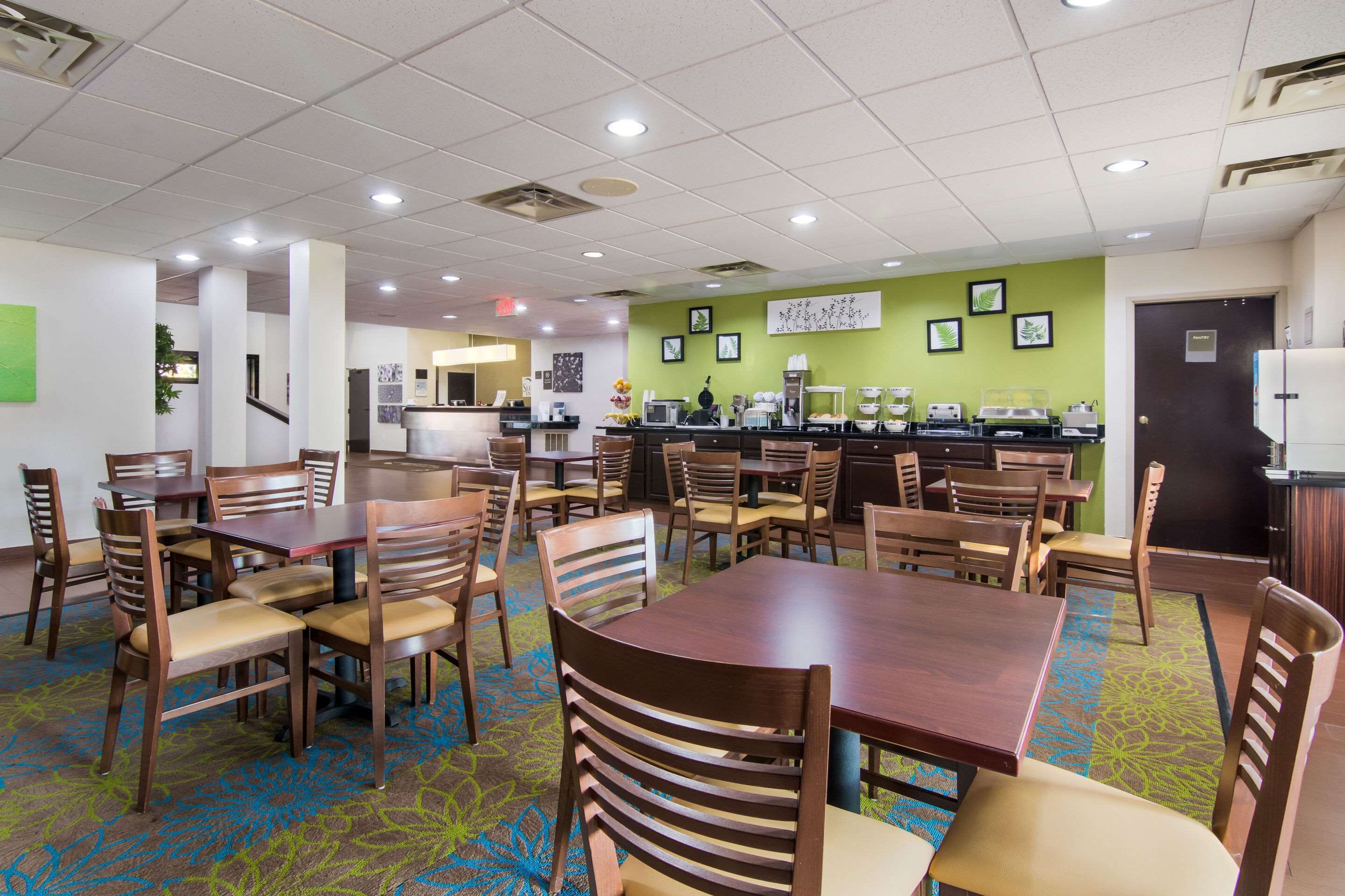 A hotel dining area with multiple tables and chairs, a coffee station, and a green accent wall with framed pictures.