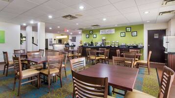 A hotel dining area with multiple tables and chairs, a coffee station, and a green accent wall with framed pictures.