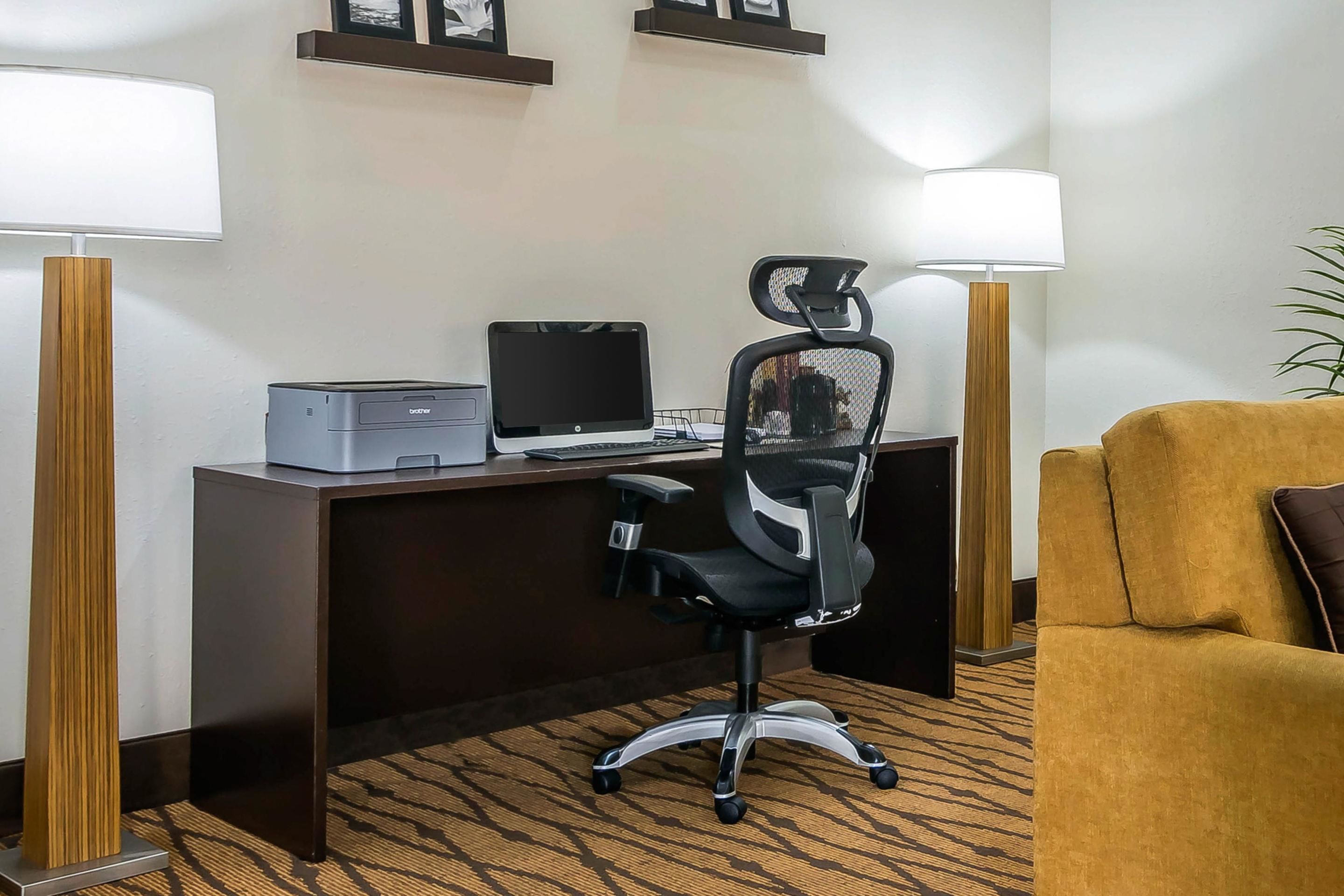 A hotel room with a desk, a computer, a printer, a black office chair, a yellow armchair, and two standing lamps.