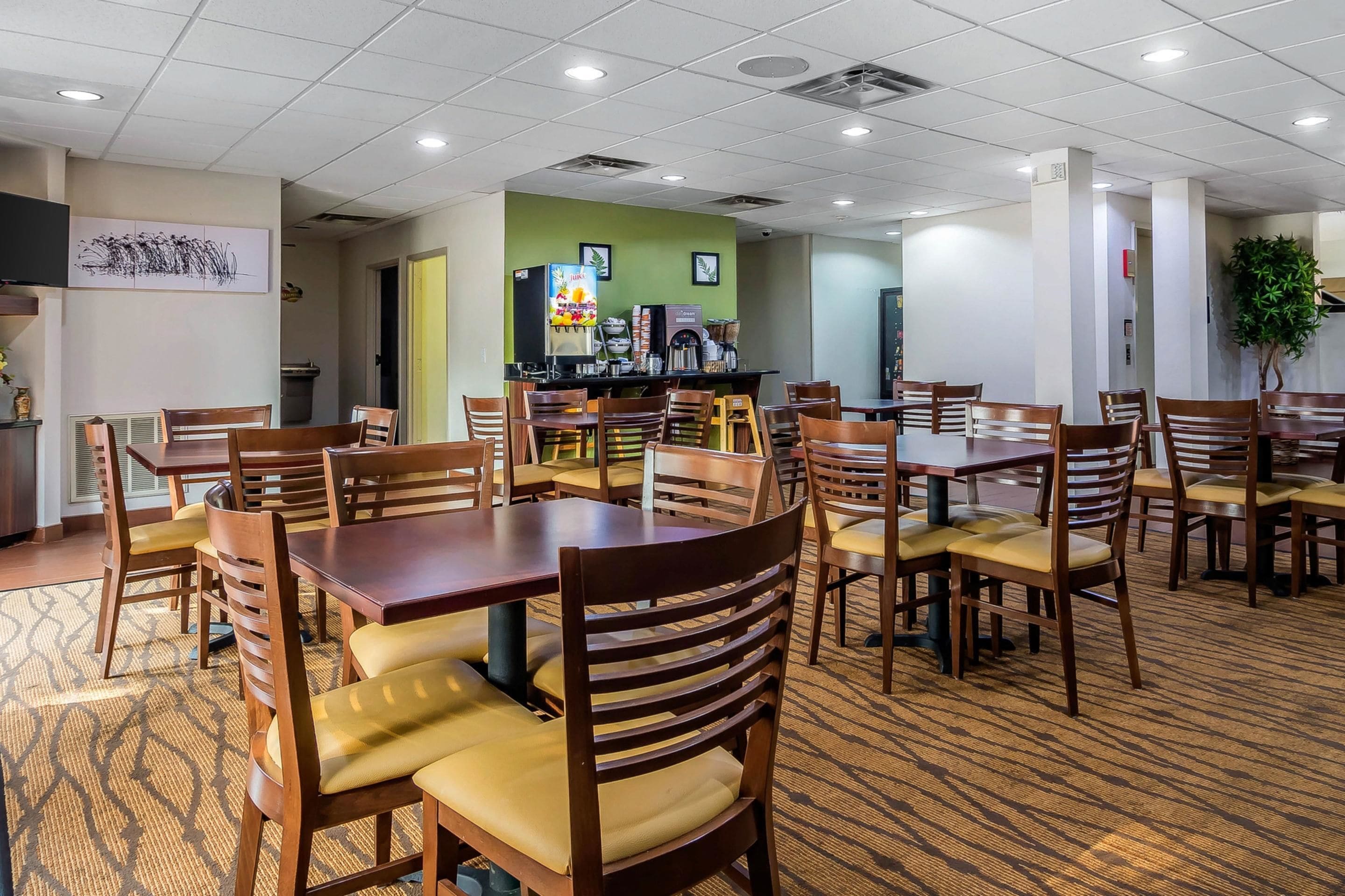 A hotel dining area with multiple wooden tables and chairs, a coffee station, and a green accent wall.
