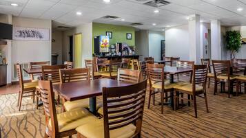 A hotel dining area with multiple wooden tables and chairs, a coffee station, and a green accent wall.
