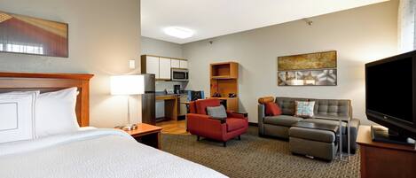 A hotel room with a large bed, a TV, a desk with a computer, a red armchair, a gray sofa, and a microwave in the kitchenette.