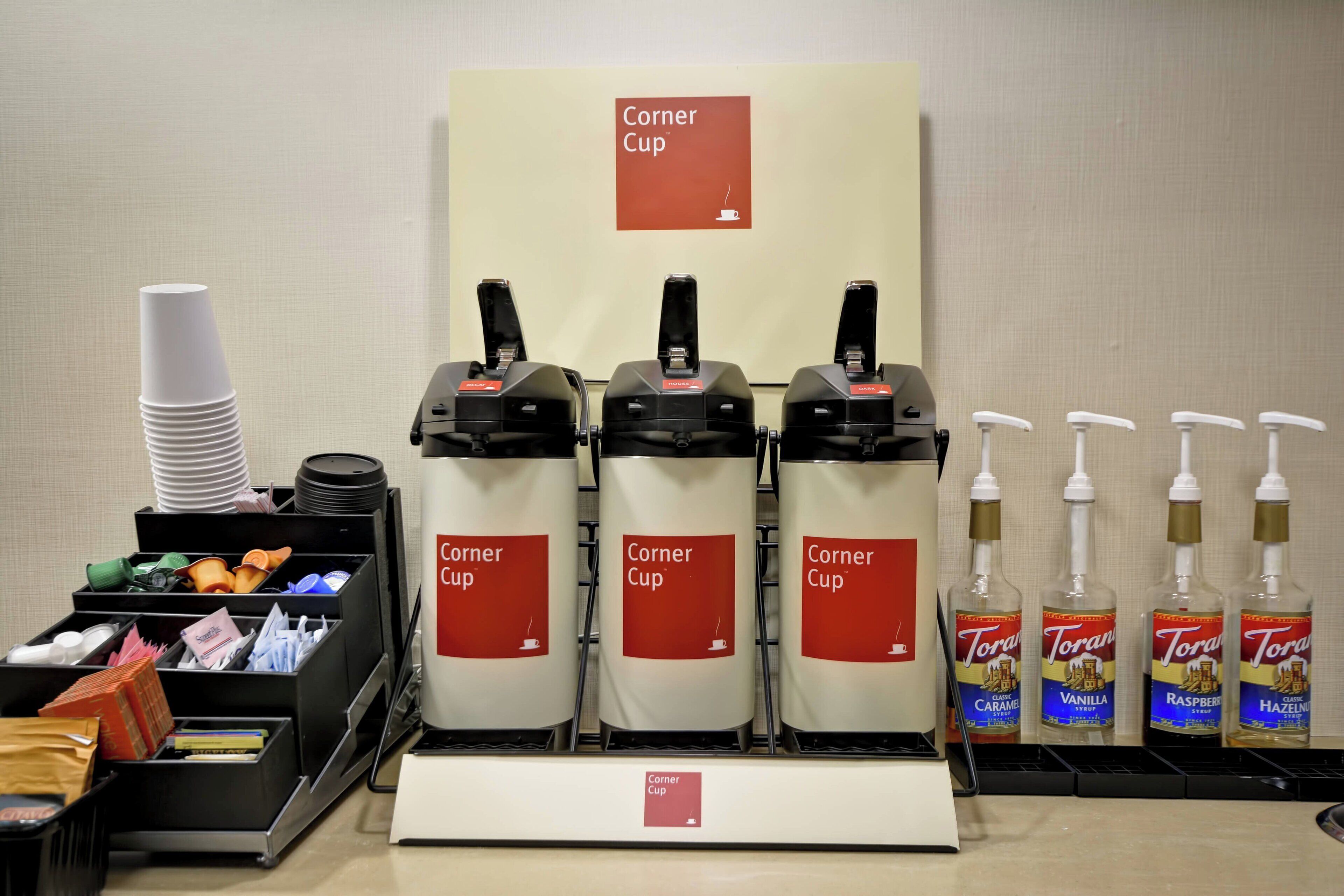 Eine Selbstbedienungs-Kaffeestation mit mehreren Spendern, beschriftet mit „Corner Cup“ und Torani-Aromasirupen.